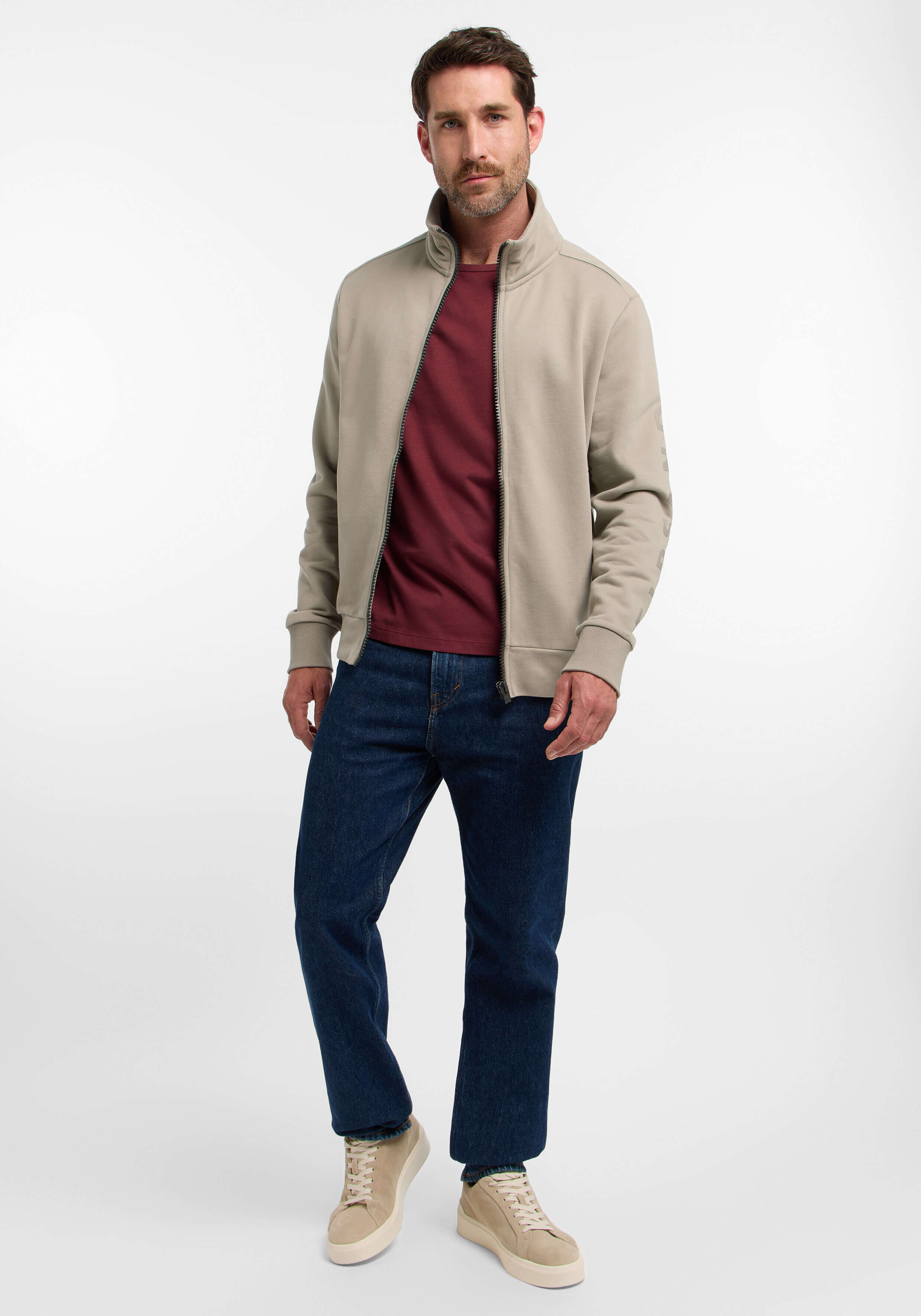 Mann trägt ein beiges Sweatshirt mit Reißverschluss, ein burgunderfarbenes T-Shirt und blaue Jeans.