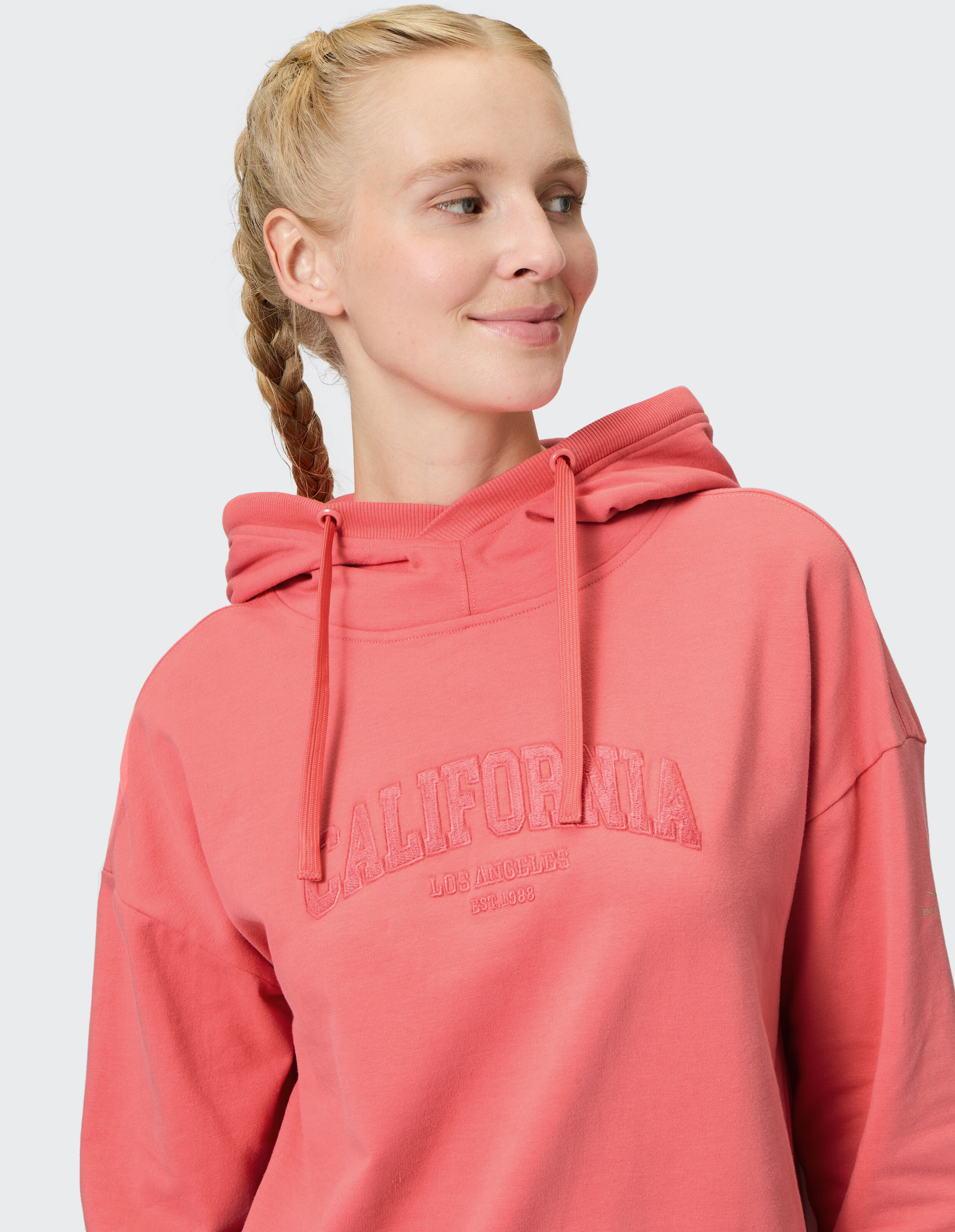 Frau trägt einen gemütlichen korallenroten Venice Beach Hoodie Muriel mit „California“-Schriftzug.