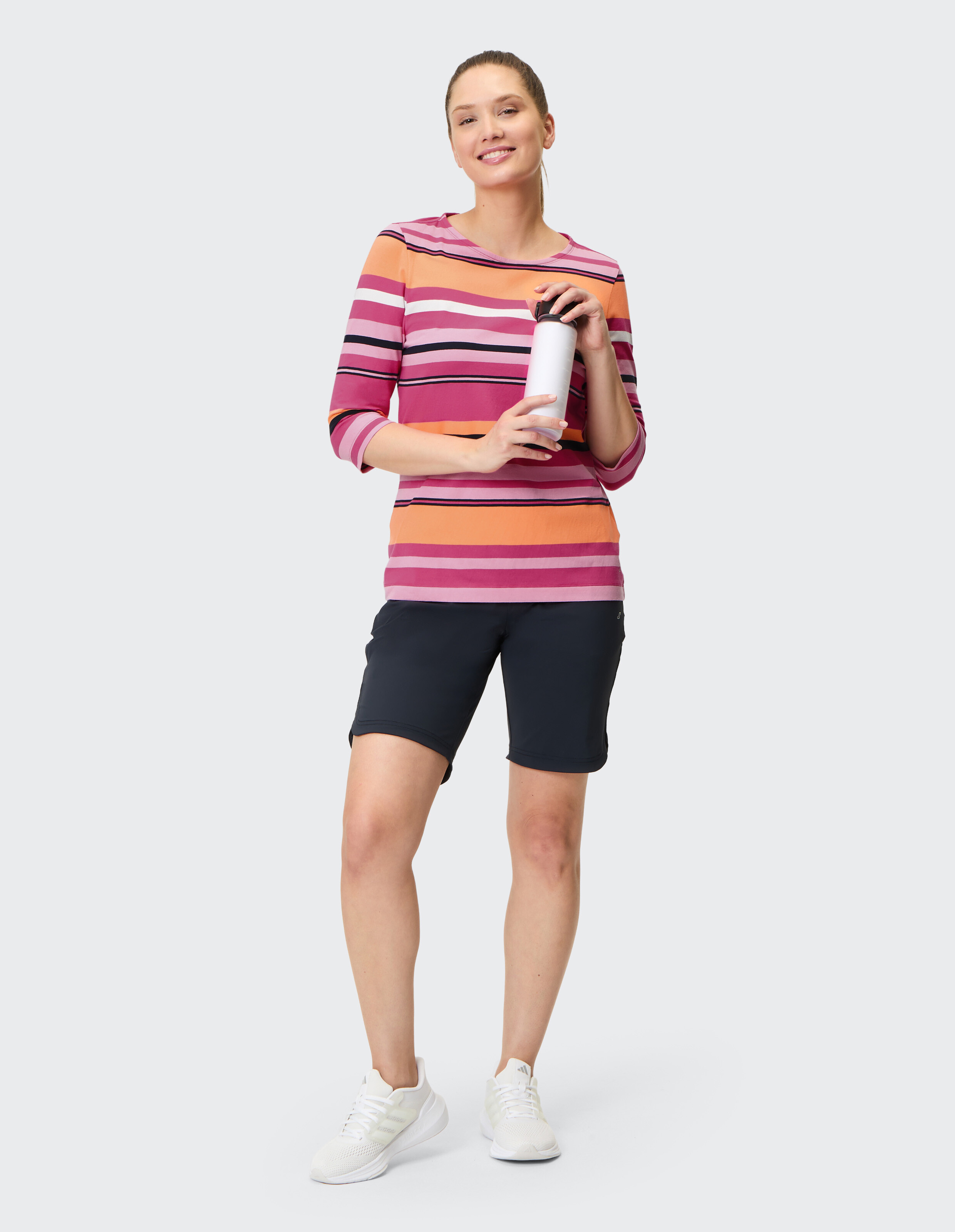 Frau in gestreiftem Pulli und schwarzer Shorts, hält eine weiße Flasche und lächelt, trägt weiße Sneaker, vor schlichtem Hintergrund.