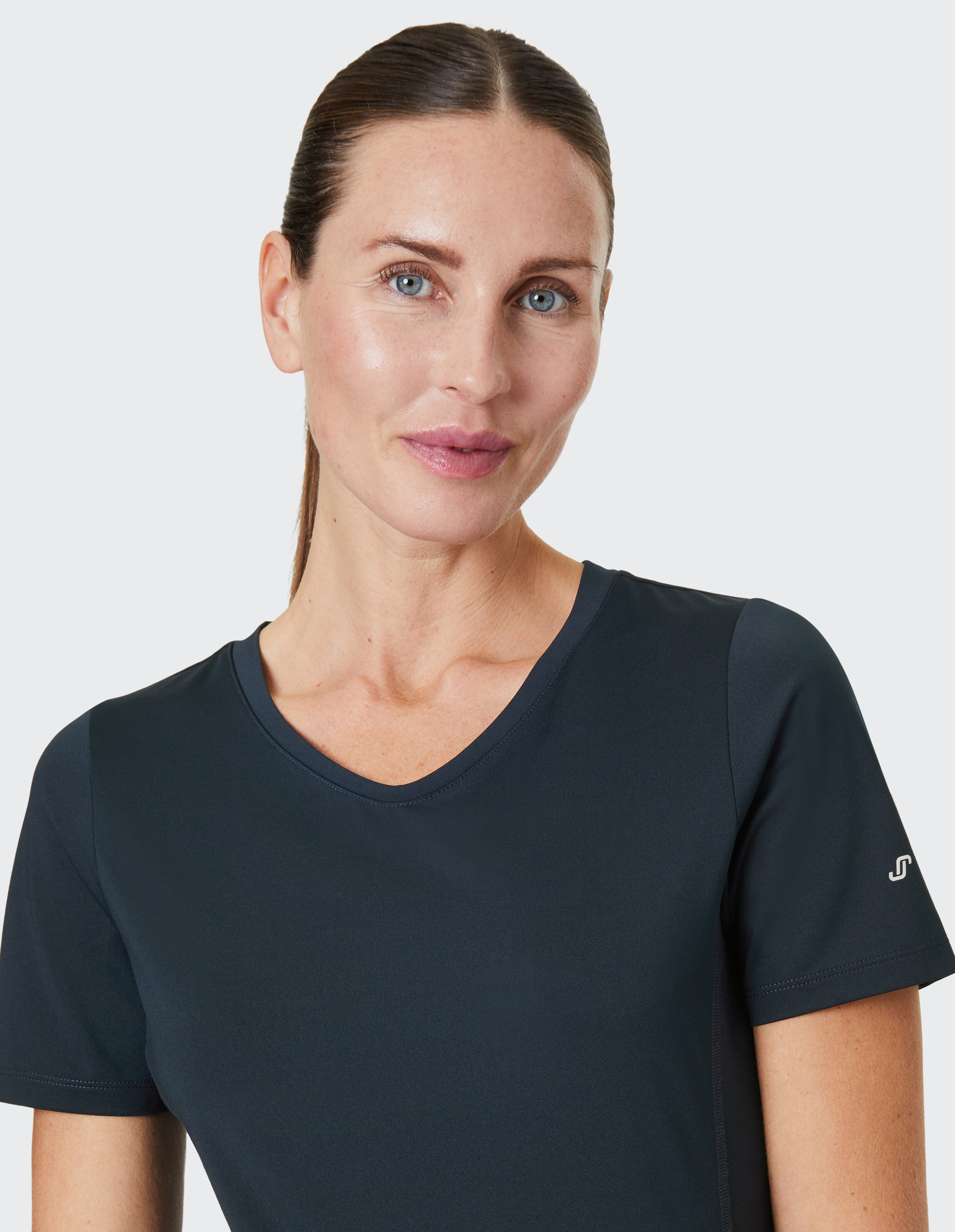 Person mit glattem braunen Haar trägt ein dunkelblaues Sportshirt und schaut lächelnd in die Kamera vor einem hellen Hintergrund.
