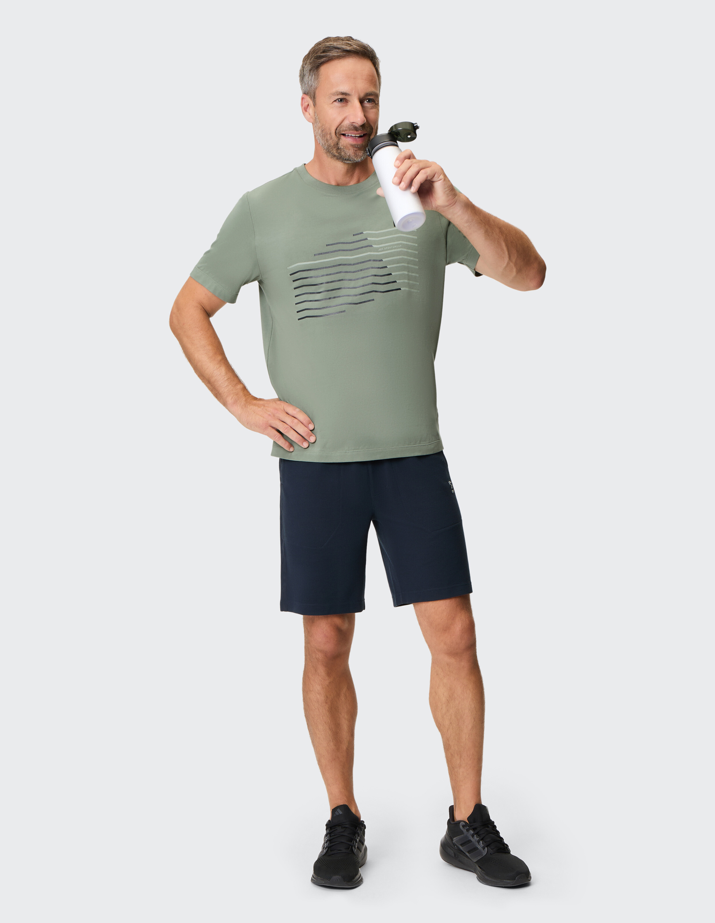 Mann in olivgrünem T-Shirt und dunkelblauen Shorts, ein Getränk in der Hand, lässiger Stil.