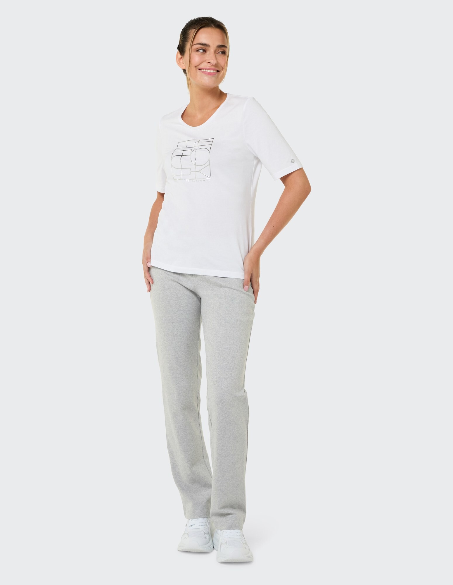 Frau in weißem Grafik-T-Shirt und grauer Jogginghose lächelt lässig.