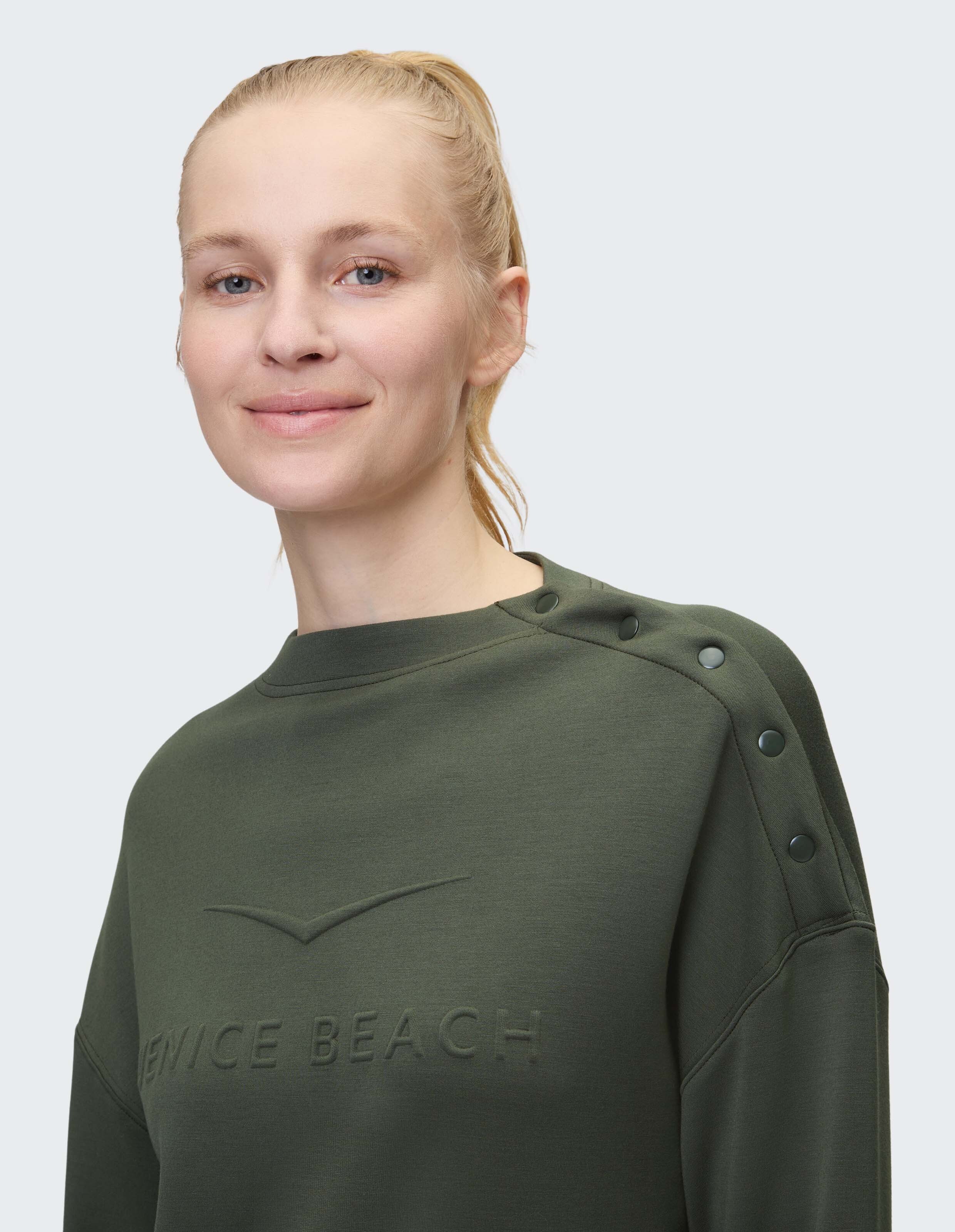 Frau trägt olivgrünes Venice Beach Cody-Sweatshirt mit Druckknöpfen an der Schulter.