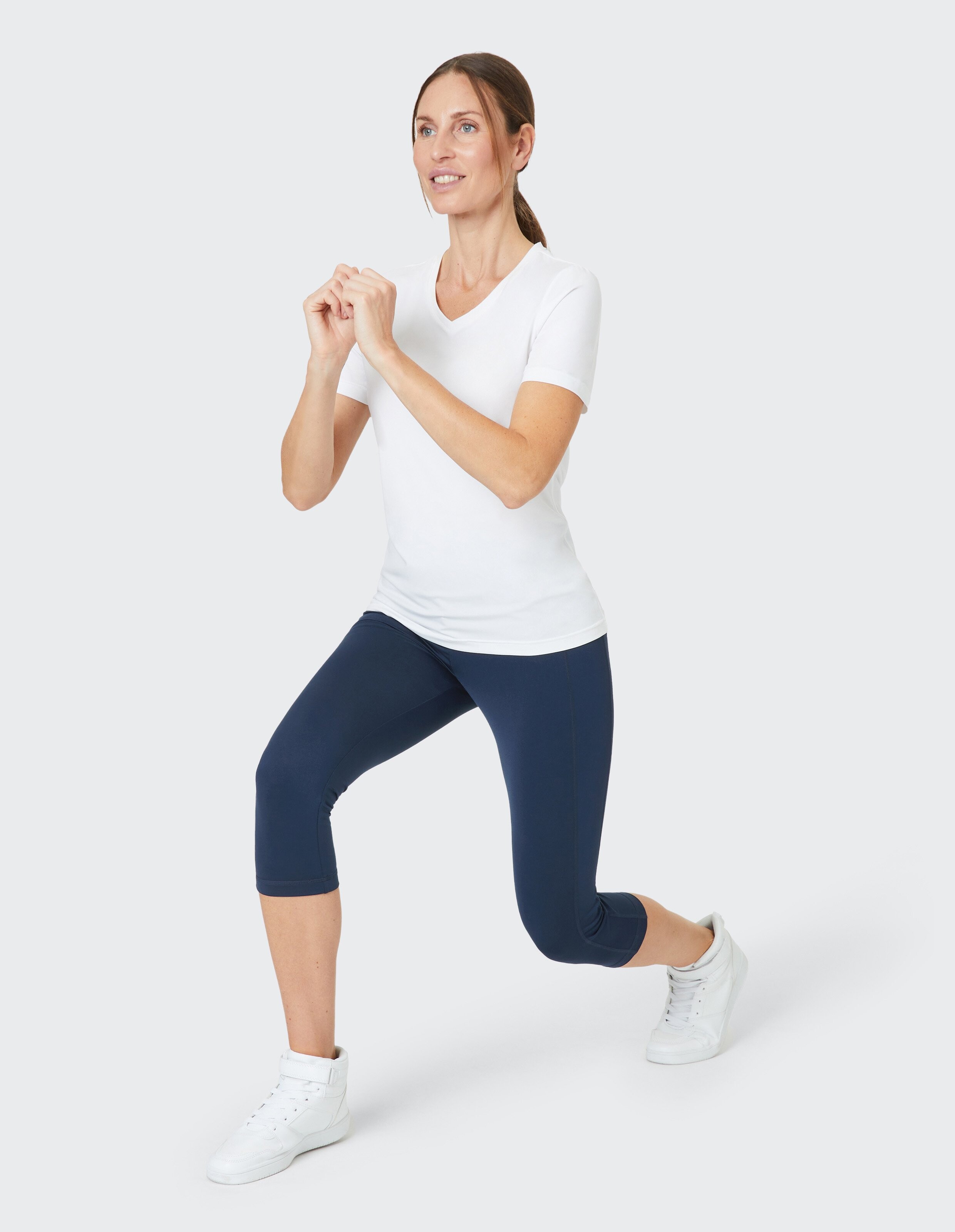Frau in weißem T-Shirt und blauen Leggings macht Lunge-Übung, Blick nach vorn, Hände zusammen, auf hellgrauem Hintergrund.