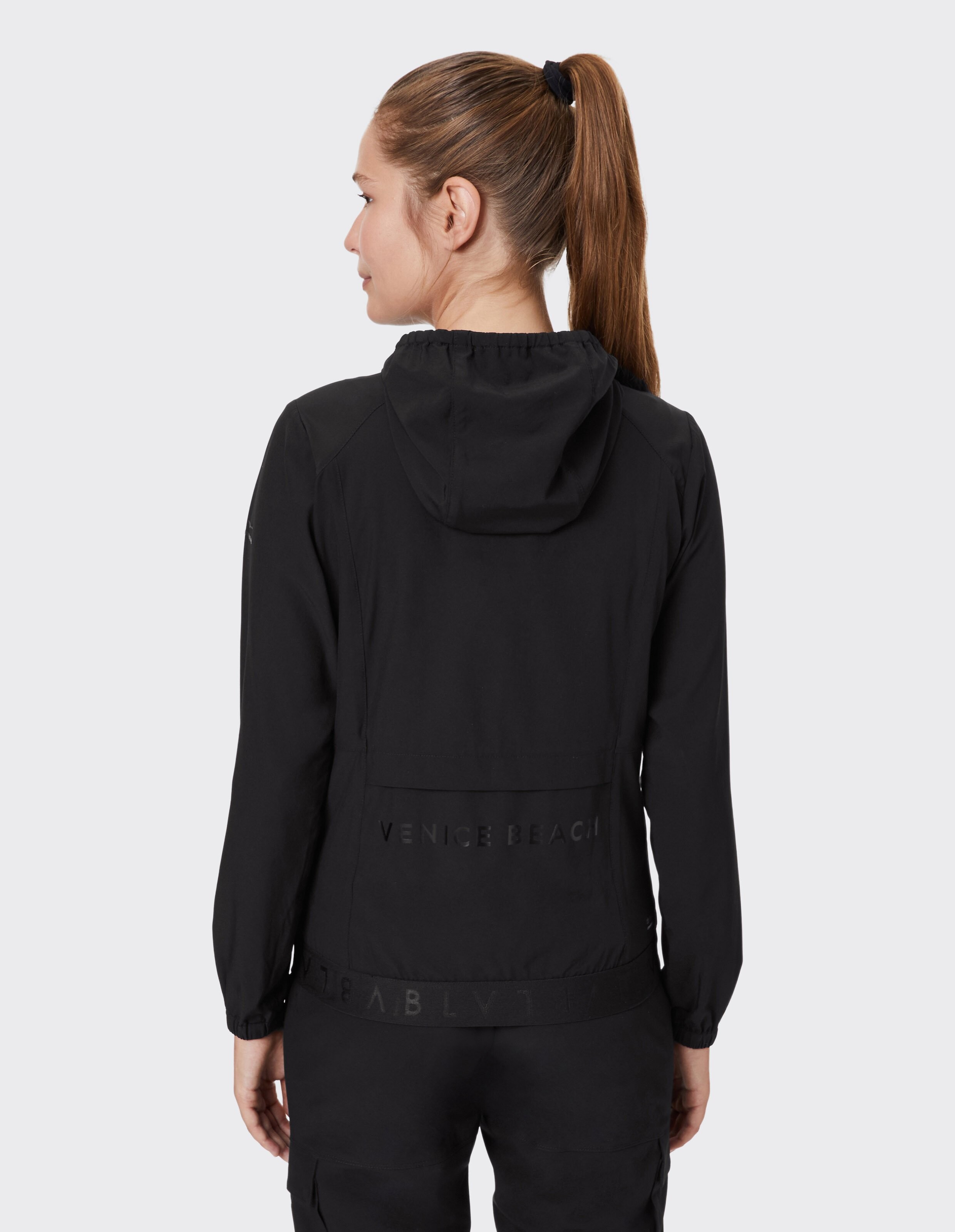 Frau trägt eine schwarze Freizeitjacke mit Kapuze und Venice Beach-Branding auf der Rückseite.