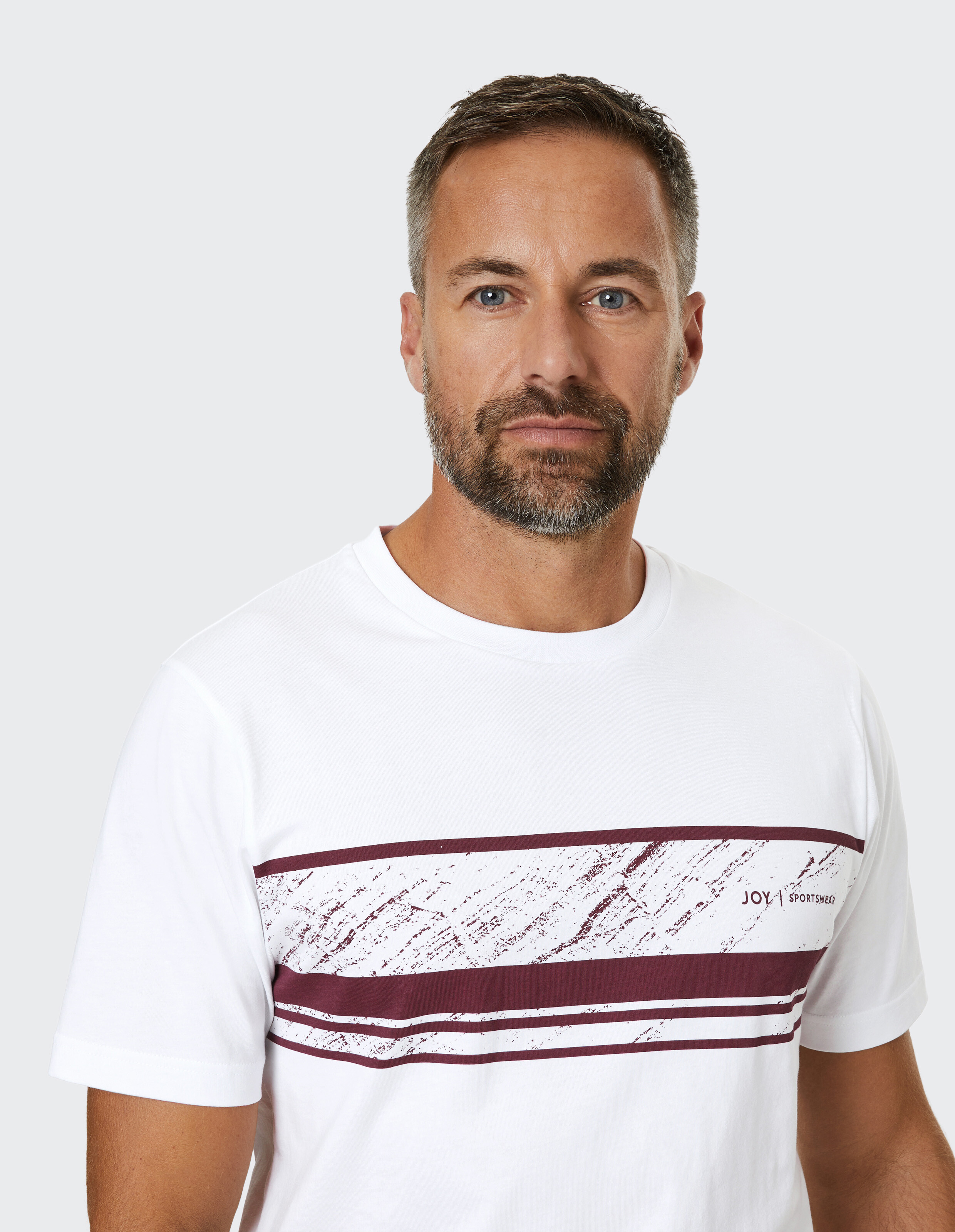 Mann mit kurzem Haar und Bart trägt ein weißes T-Shirt mit weinroten Streifen, vor hellem Hintergrund.