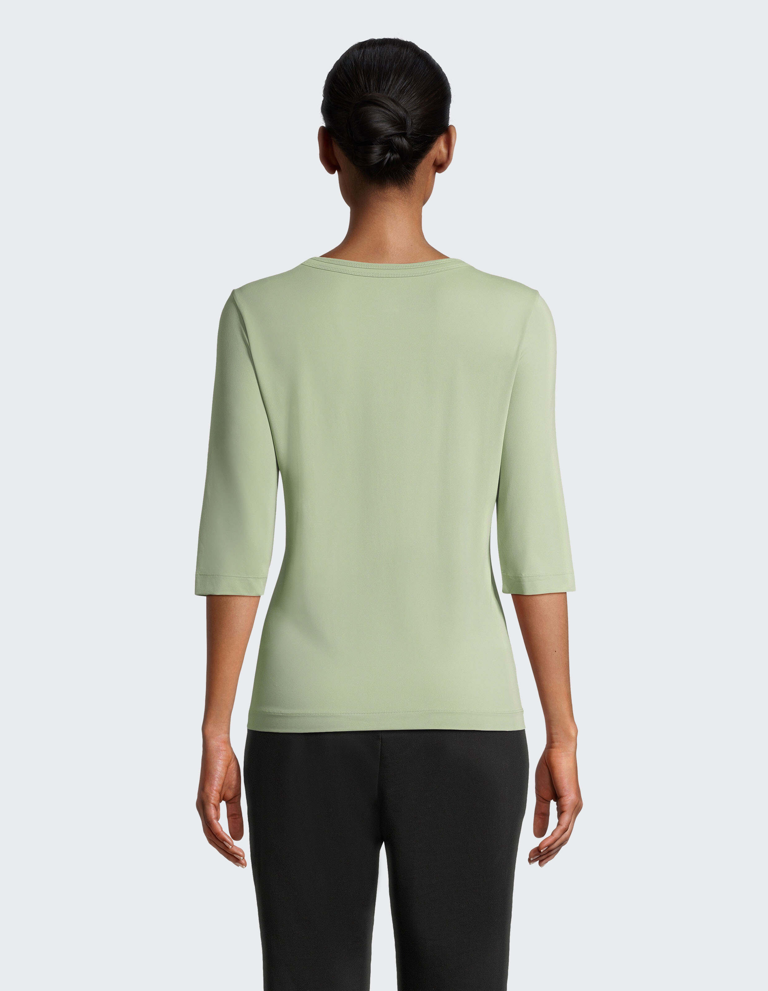 Person von hinten in einem hellgrünen 3/4-Arm-Shirt und schwarzer Hose, Haar in einem eleganten Knoten, vor grauem Hintergrund.