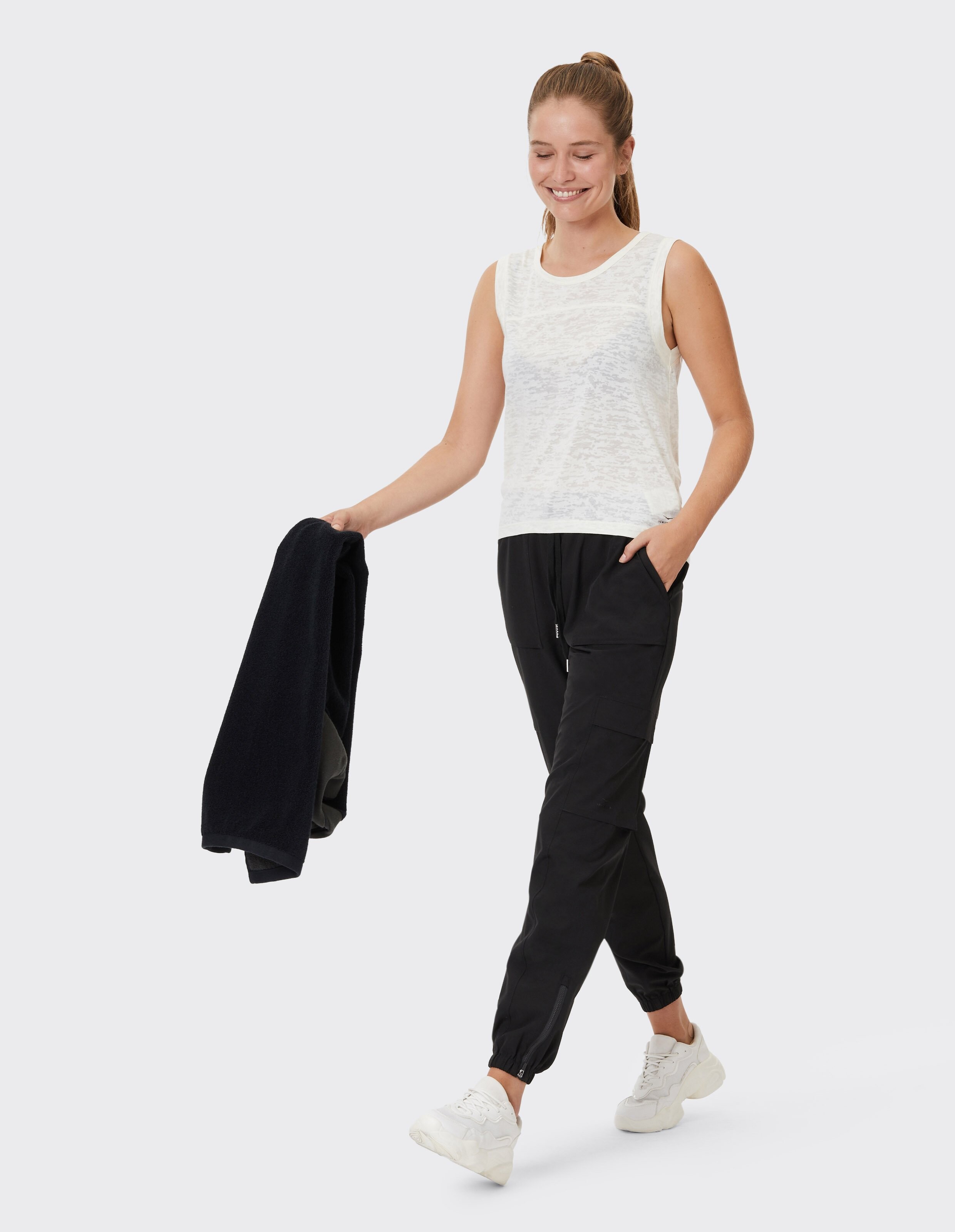 Frau trägt ein weißes Tanktop und schwarze lässige Jogginghosen und hält eine schwarze Fleecejacke in der Hand.
