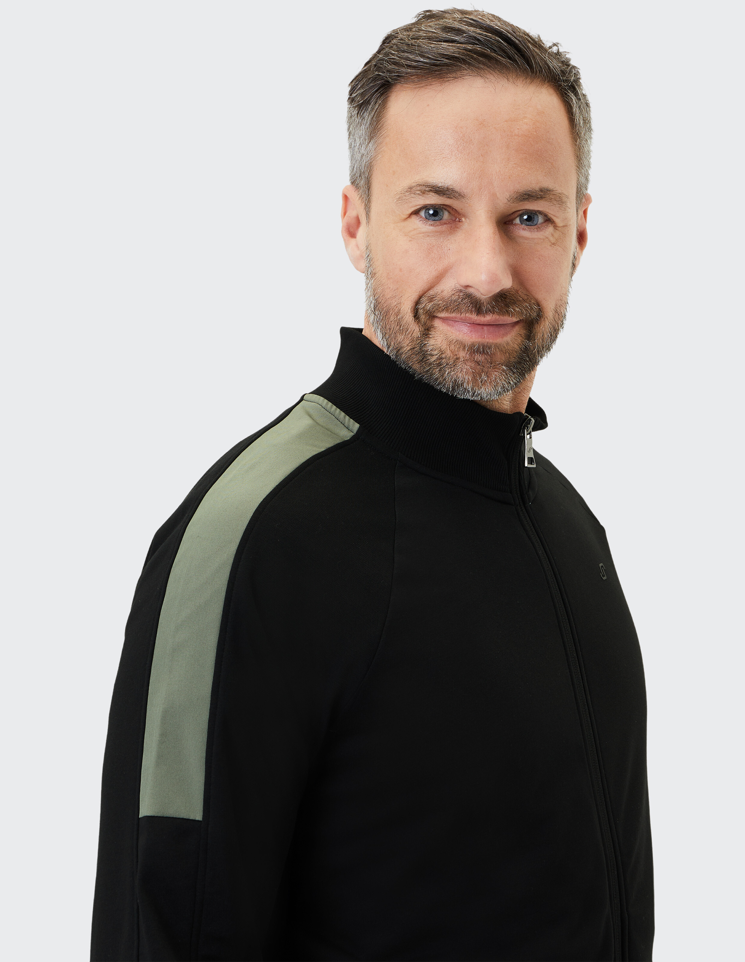 Ein lächelnder Mann mit grauem Haar und Bart trägt eine schwarze Jacke mit olivfarbenem Streifen auf der Schulter vor grauem Hintergrund.