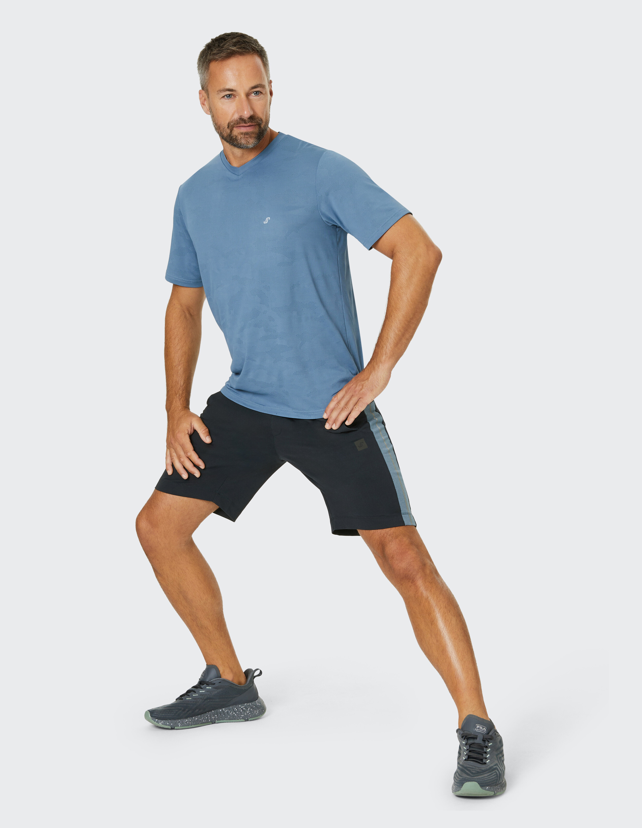 Mann in sportlicher Haltung trägt ein blaues T-Shirt, schwarze Shorts und Sneakers, posiert mit einer Hand auf der Hüfte.