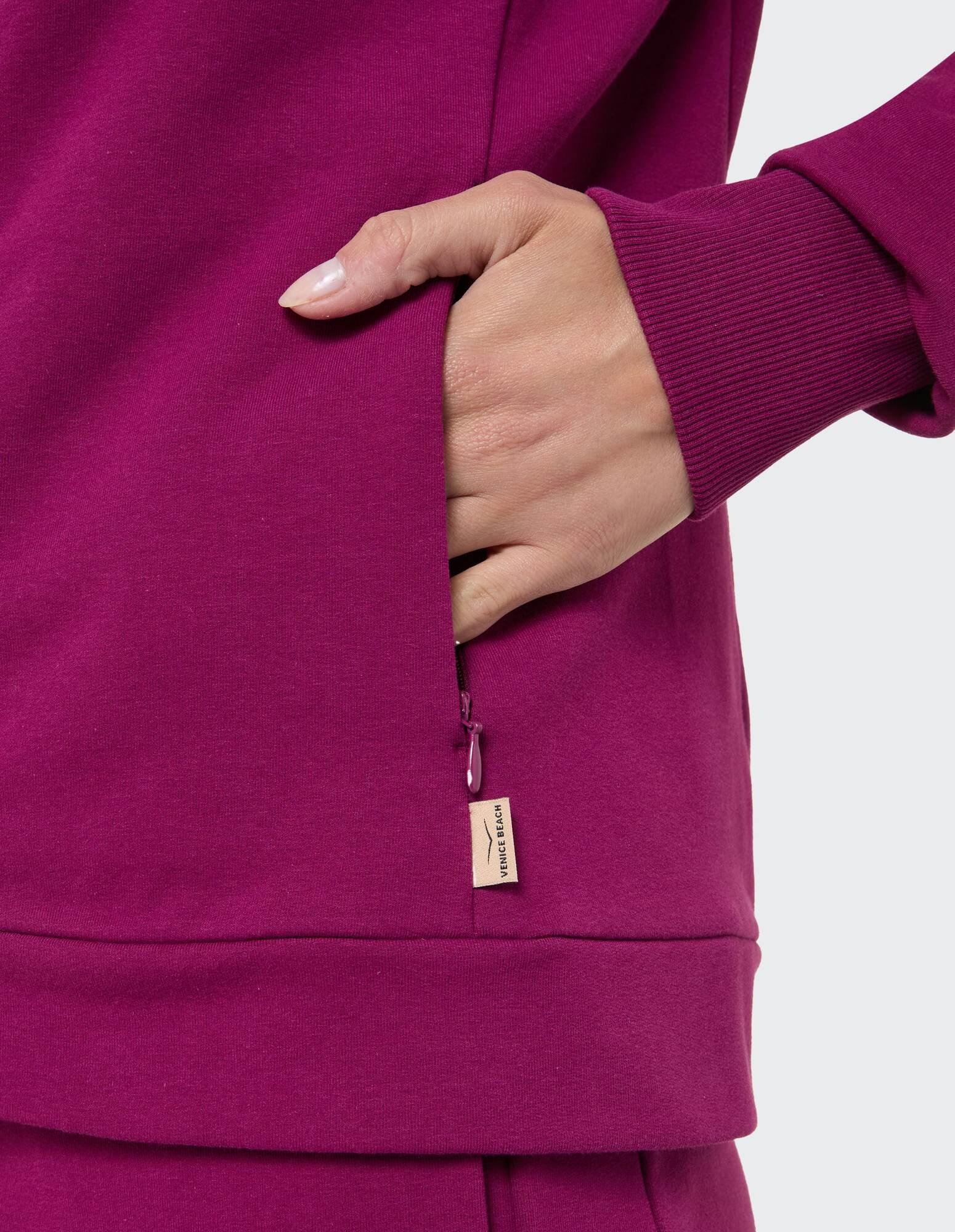 Nahaufnahme einer Hand in einer Reißverschlusstasche eines fuchsiafarbenen Freizeit-Sweatshirts.