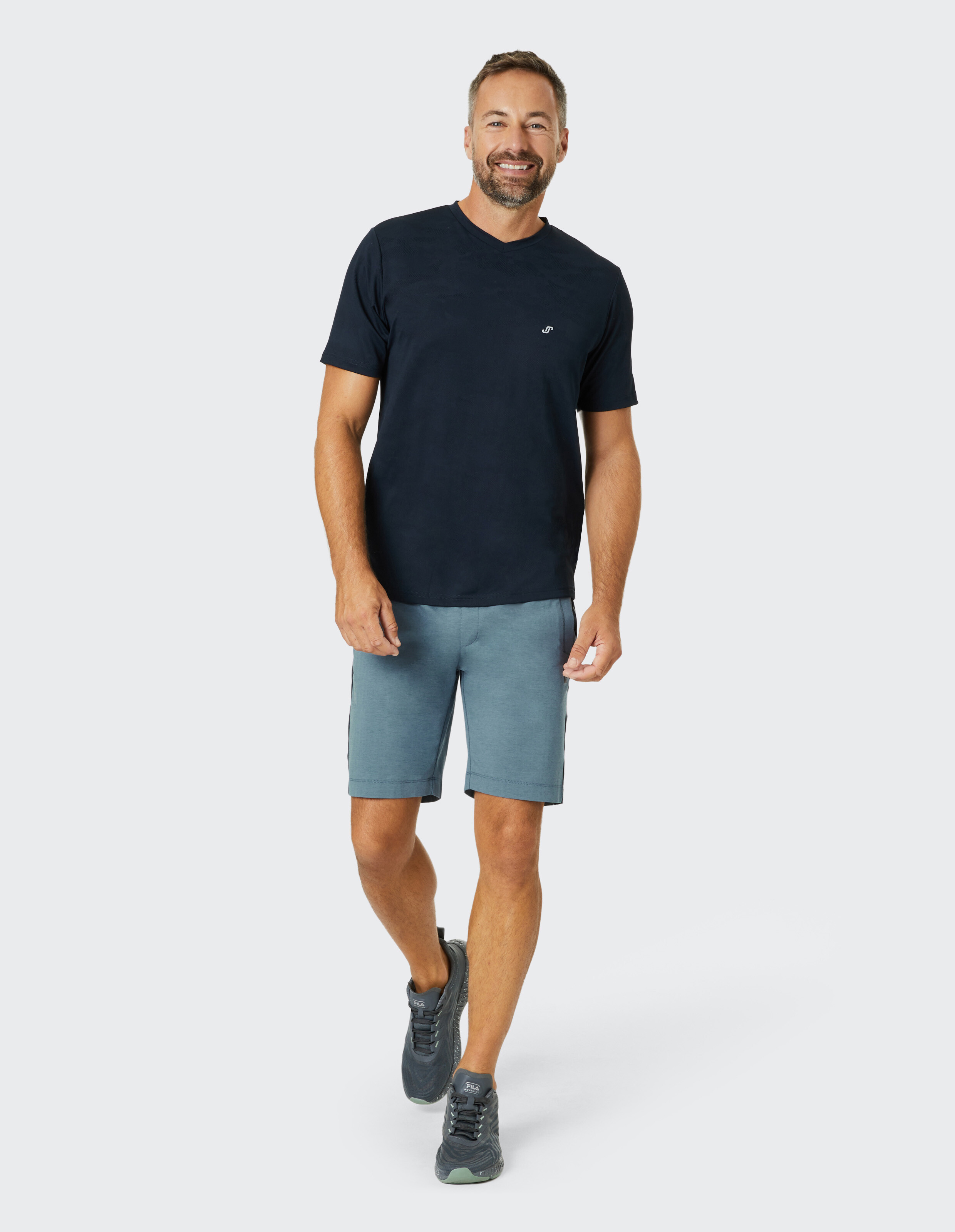Mann in dunkelblauem T-Shirt und grauen Shorts geht lächelnd auf weißem Hintergrund, trägt graue Sneakers.
