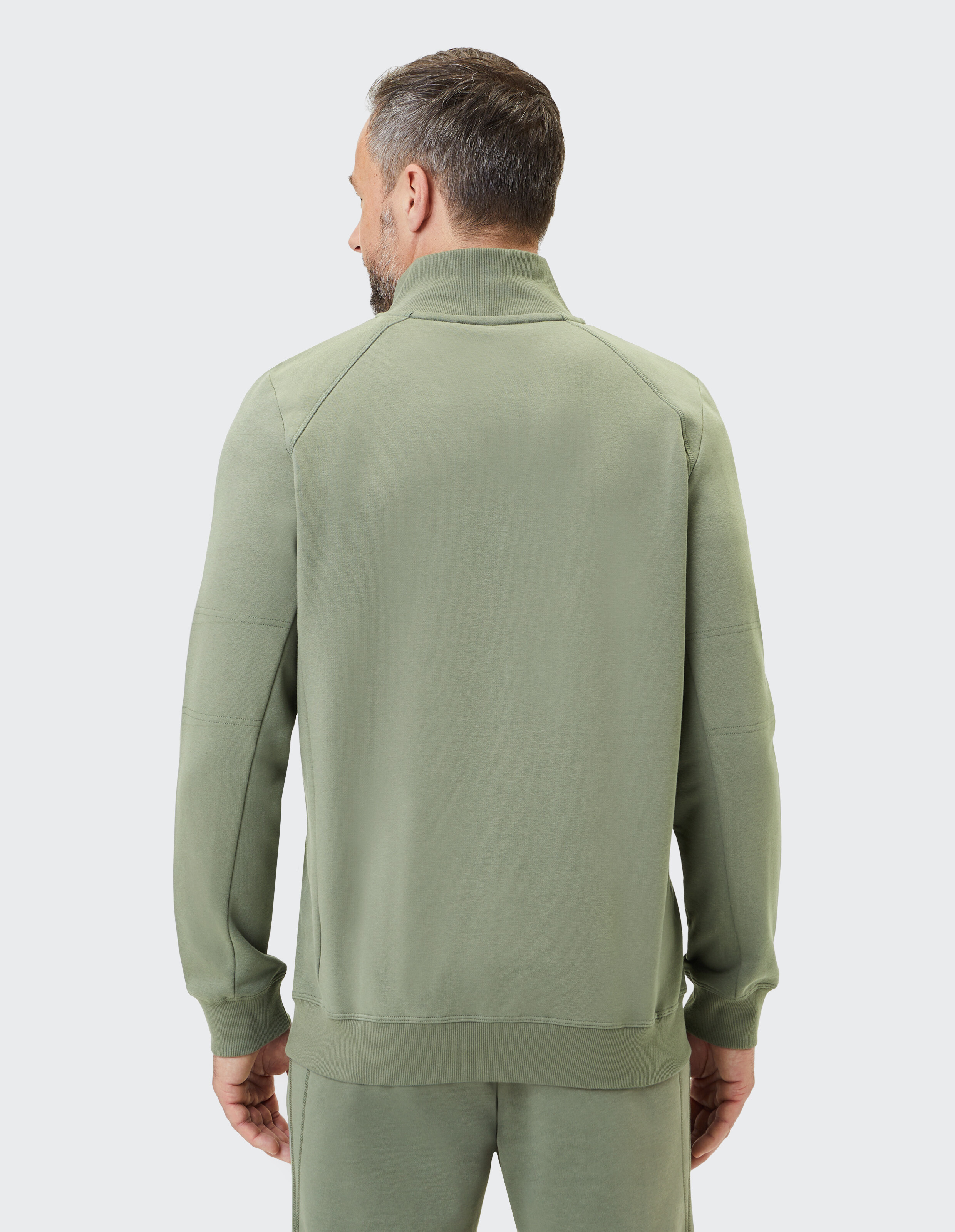 Ein Mann mit kurzem Haar trägt eine olivgrüne Trainingsjacke mit Kragen. Er steht mit dem Rücken zur Kamera vor grauem Hintergrund.