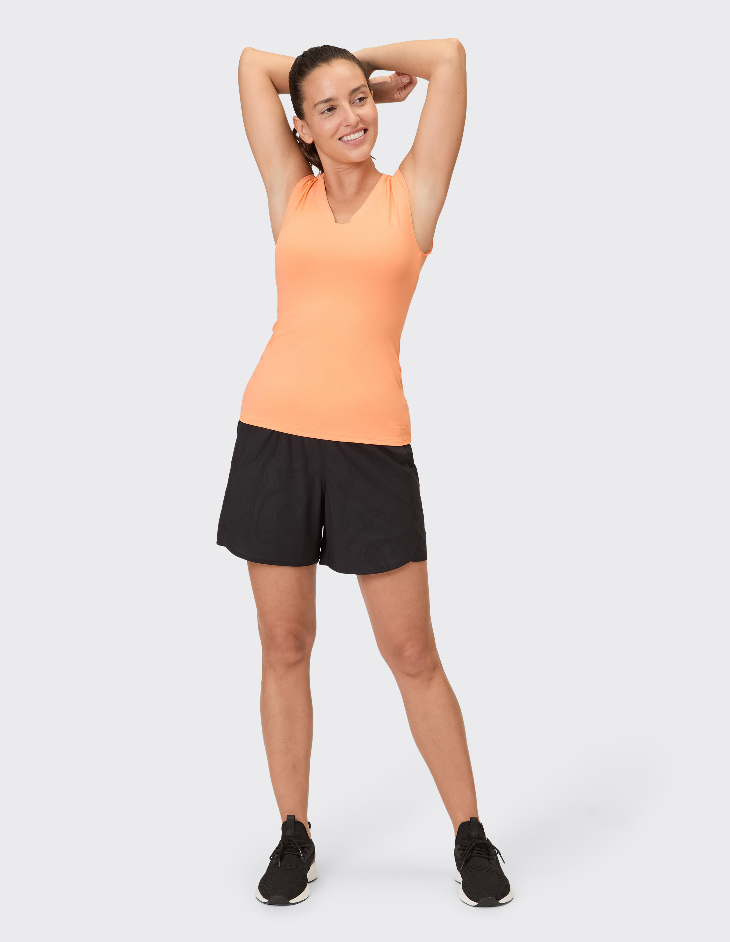 Frau in orangefarbenem Tanktop und schwarzen Shorts, lächelnd und lässig posierend.