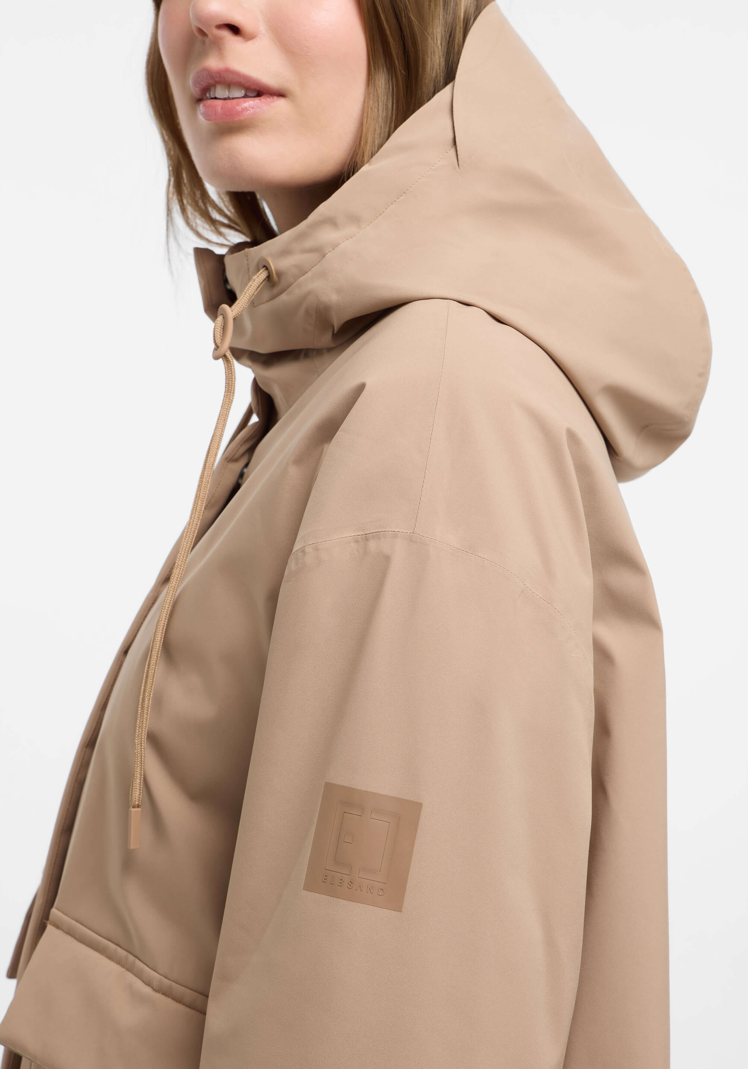 Nahaufnahme einer Frau, die eine beige Kapuzenjacke mit Logodetail trägt.