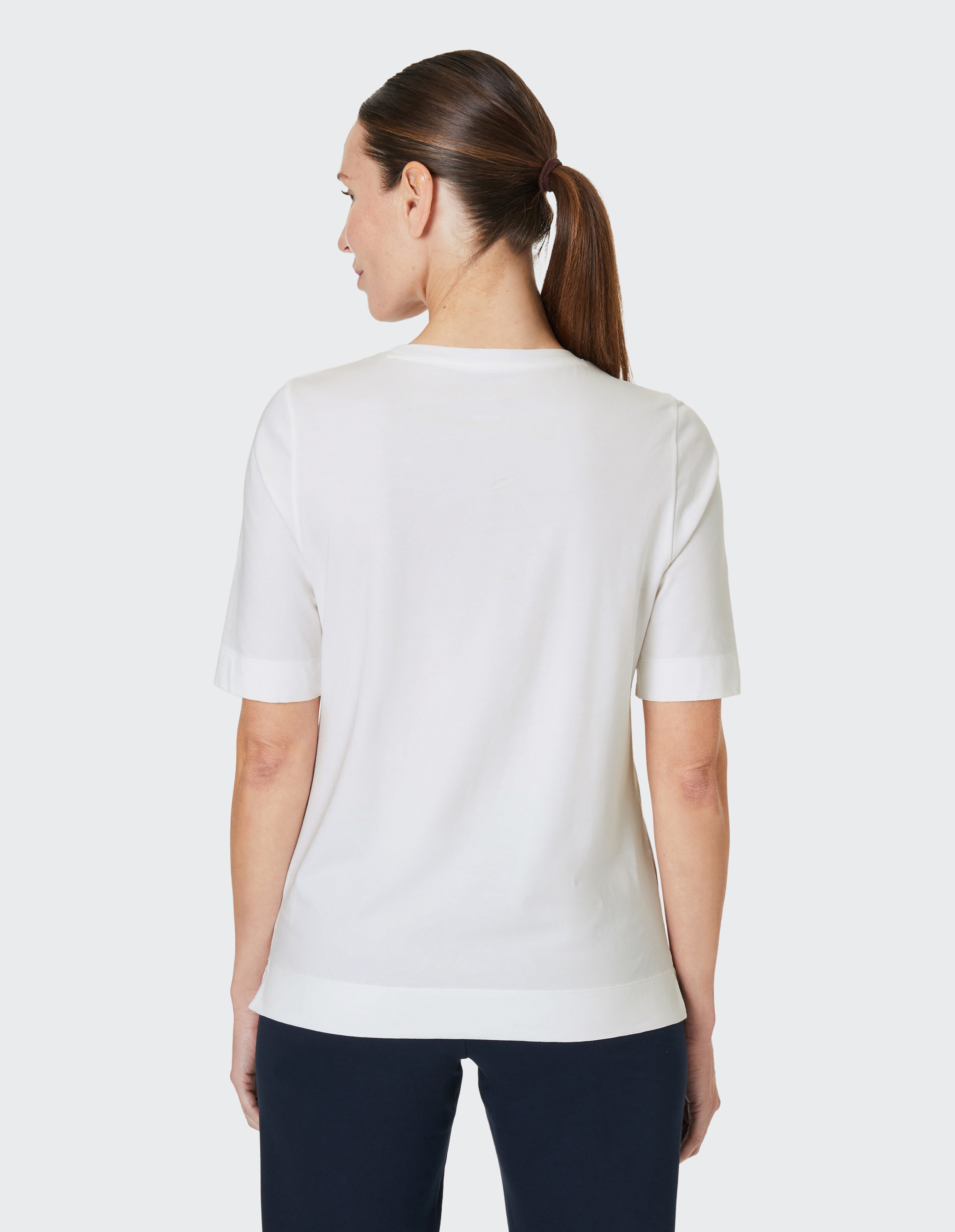 Eine Frau mit glattem, braunem Haar im Pferdeschwanz trägt ein weißes T-Shirt und dunkle Hose, ist von hinten fotografiert.
