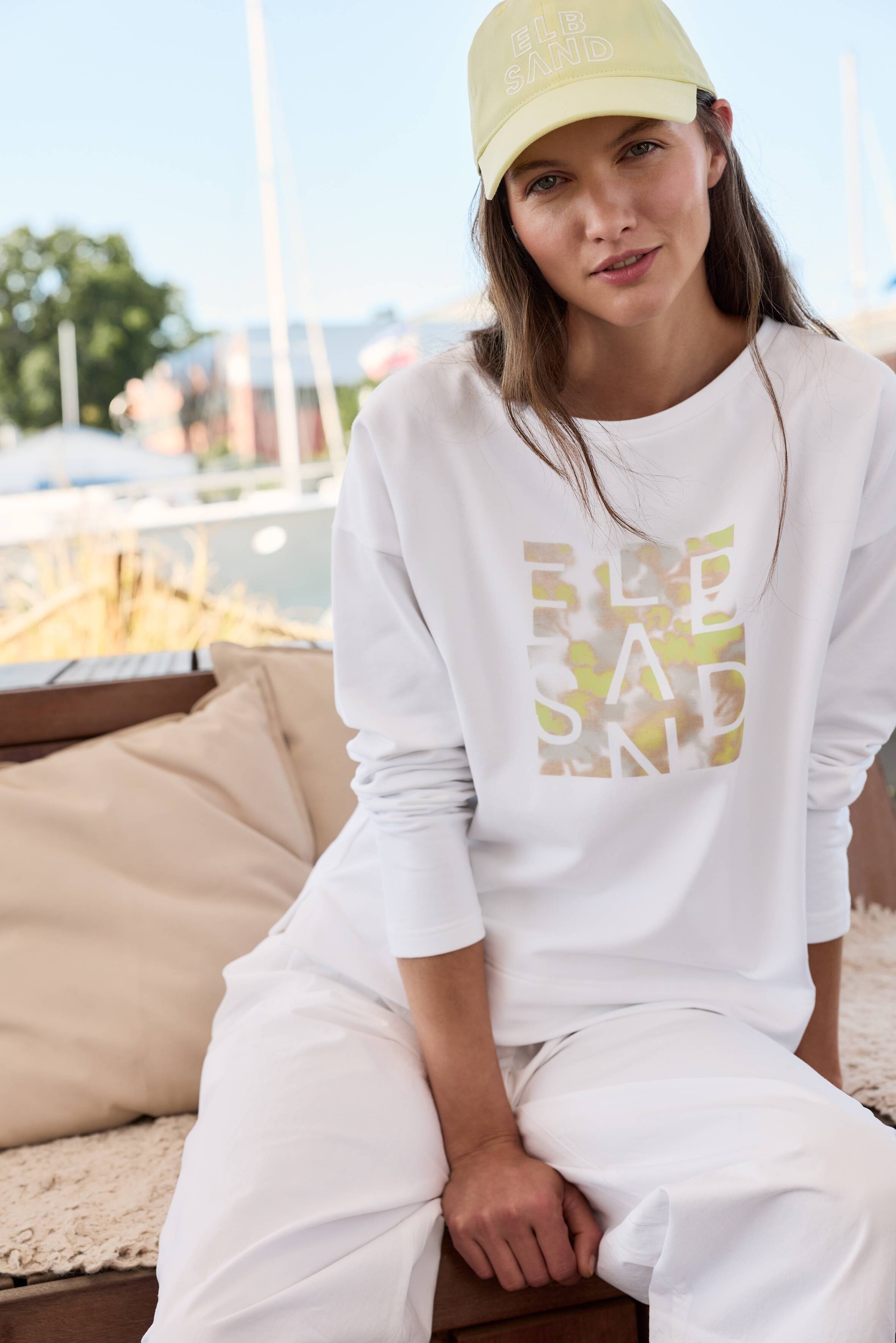 Frau im weißen ELBSAND-Sweatshirt und gelber Mütze, perfekt für gemütliche, lässige Lounge-Kleidung.