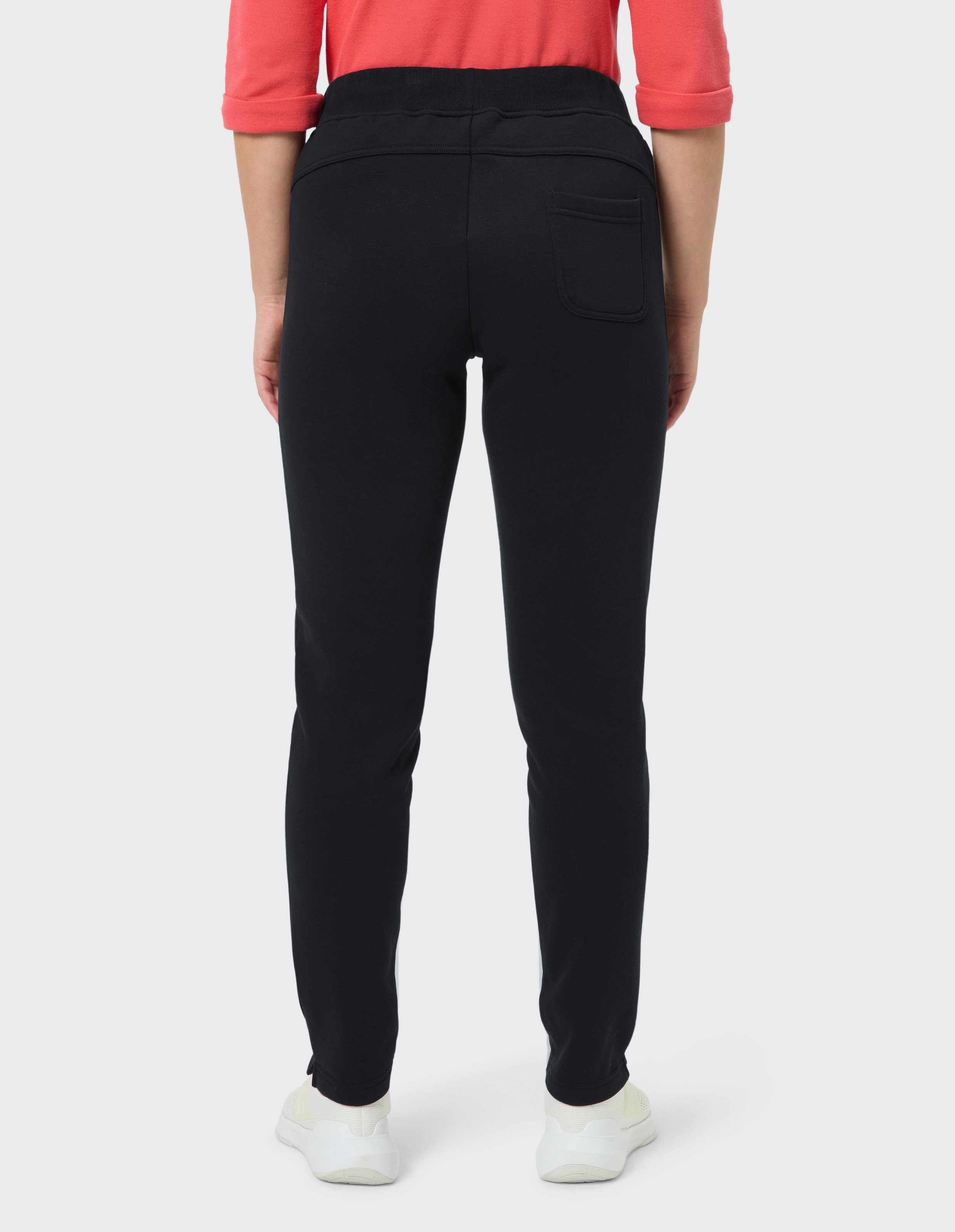 Person in einem rosa gestreiften Shirt trägt schwarze Jogginghose und hellrosa Sneakers mit rosa Sohlen, steht mit überkreuzten Beinen.