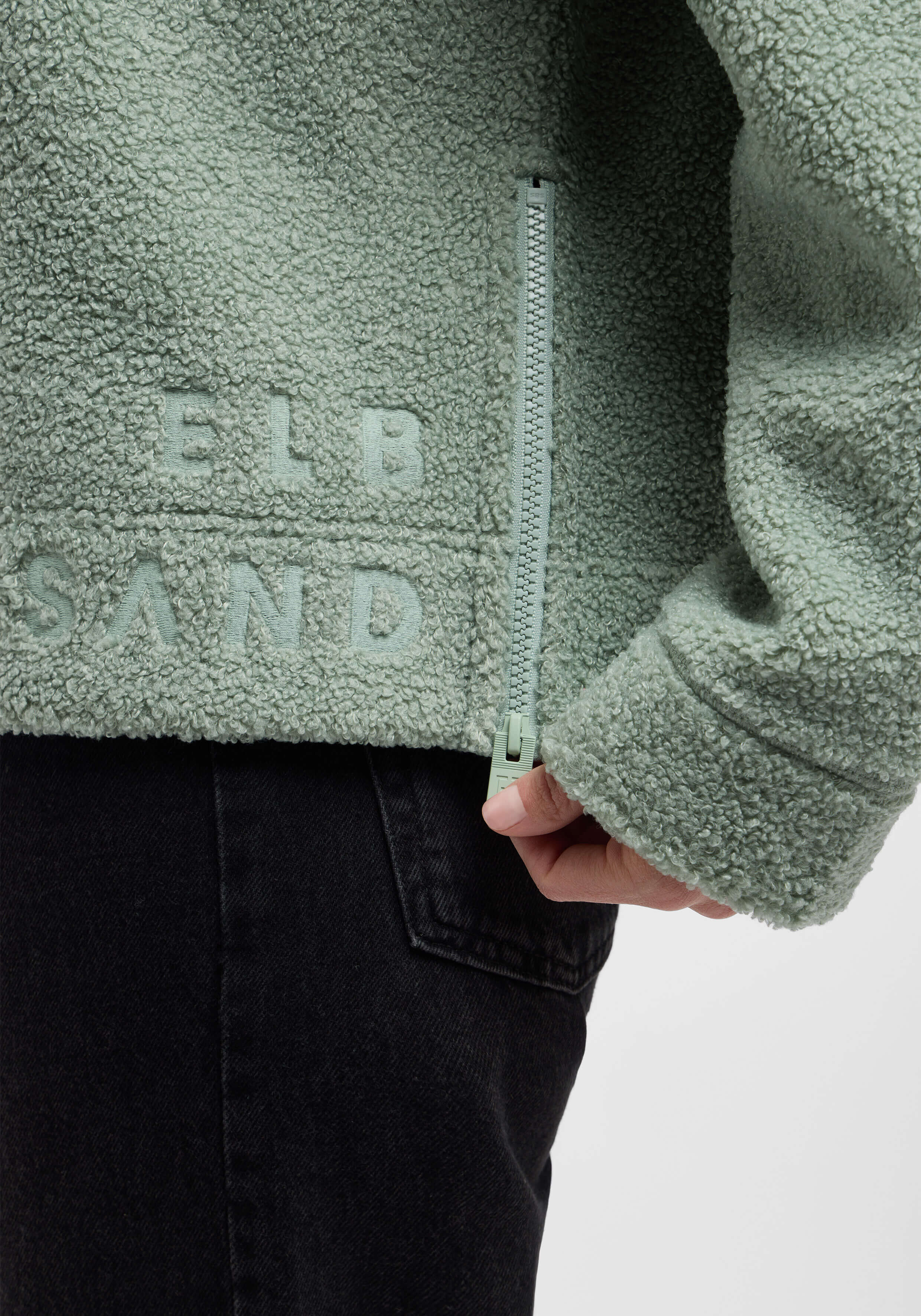 Nahaufnahme einer kuscheligen grünen Fleecejacke mit Reißverschluss und ELBSAND-Logo.