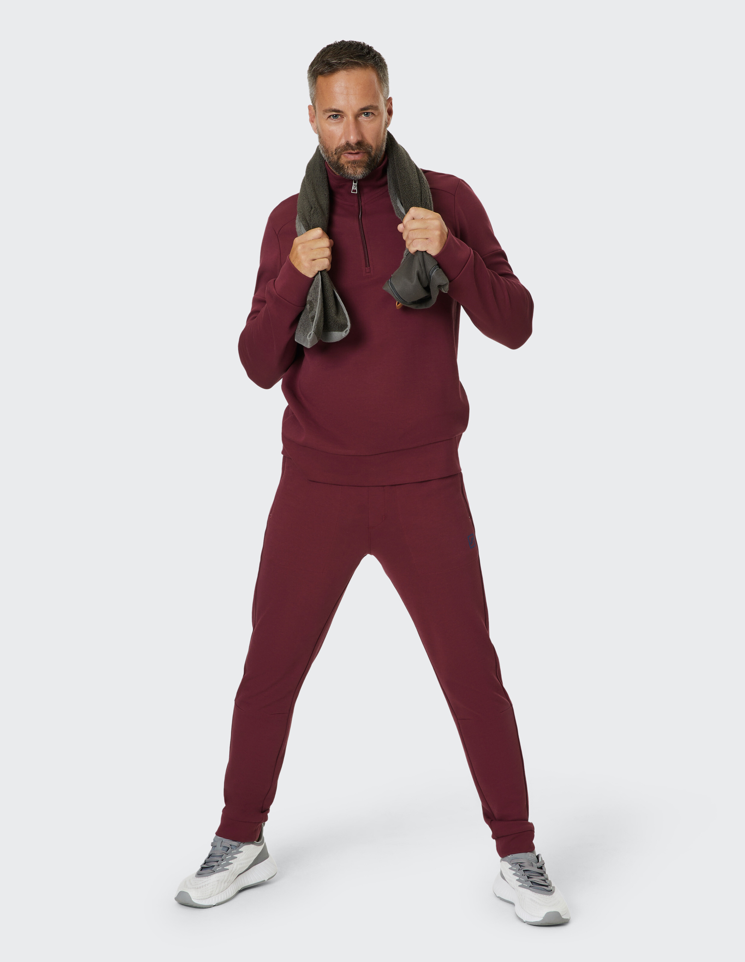 Ein Mann in burgunderfarbenem Jogginganzug steht mit gespreizten Beinen, hält ein graues Handtuch um den Nacken, weißer Hintergrund.