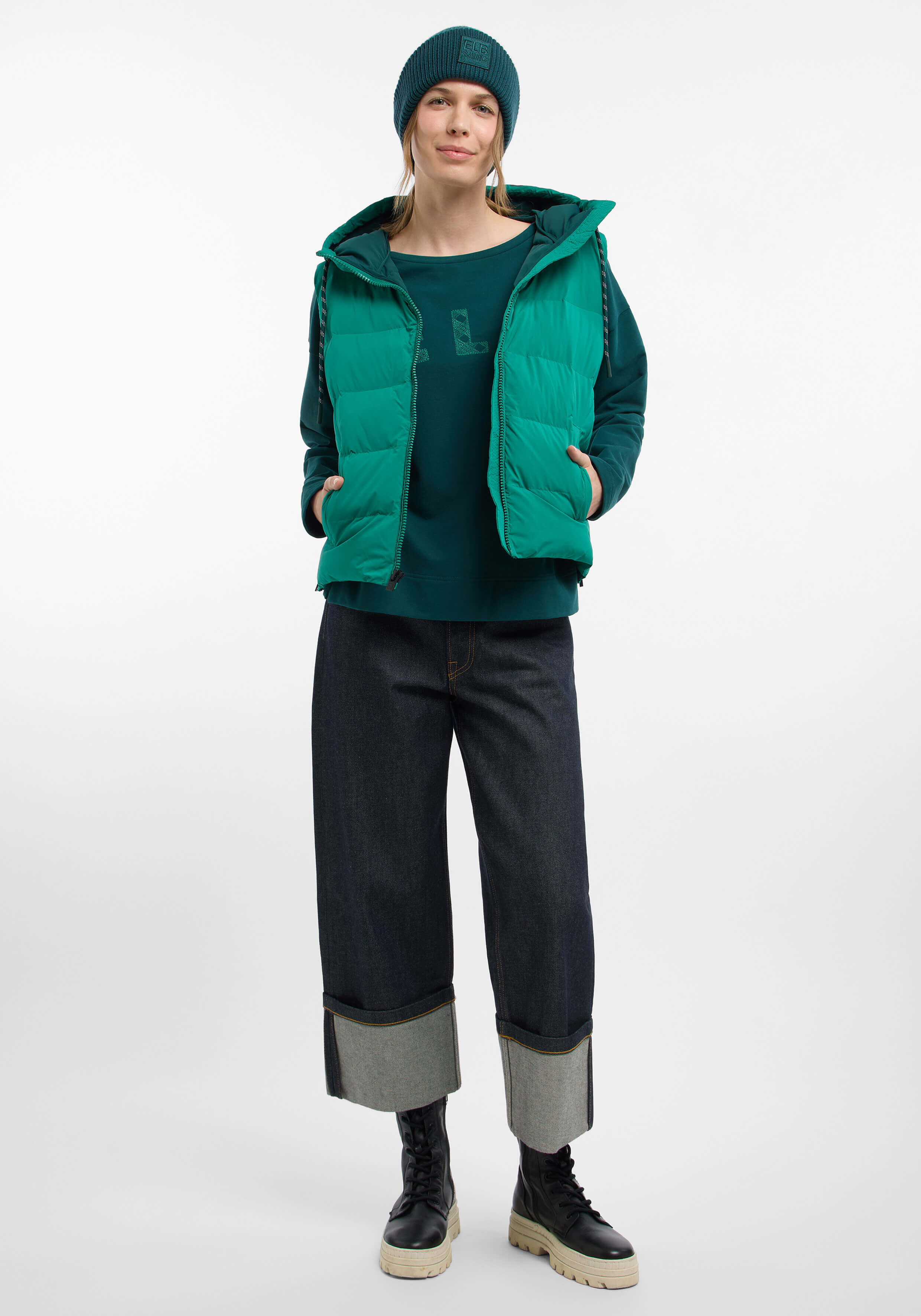 Frau trägt eine grüne Steppweste, ein Sweatshirt, eine Mütze und Jeans mit Bündchen.