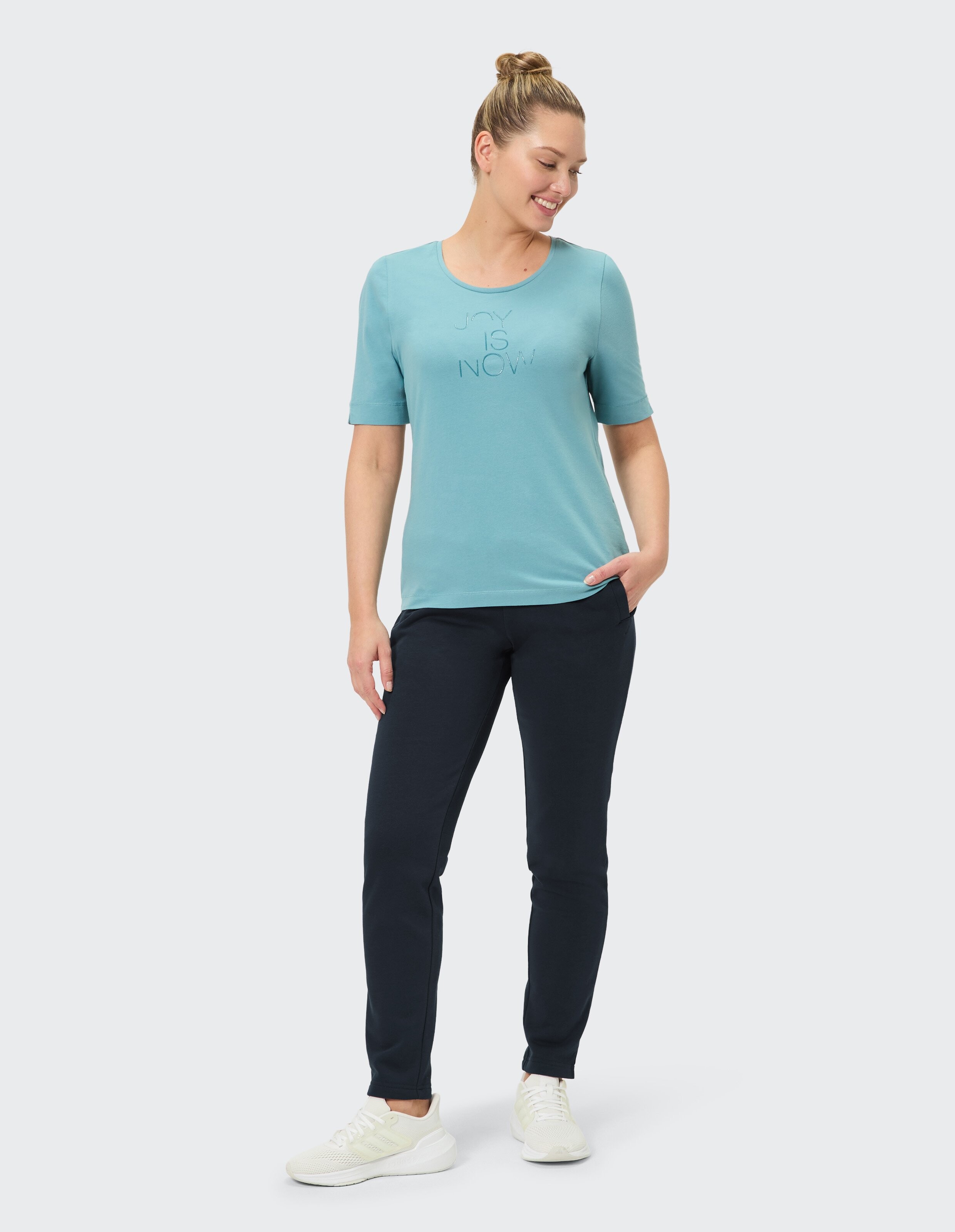 Frau in sportlicher Pose trägt ein helles T-Shirt mit der Aufschrift go with the flow und dunkle Jogginghose und rosafarbene Sneaker.