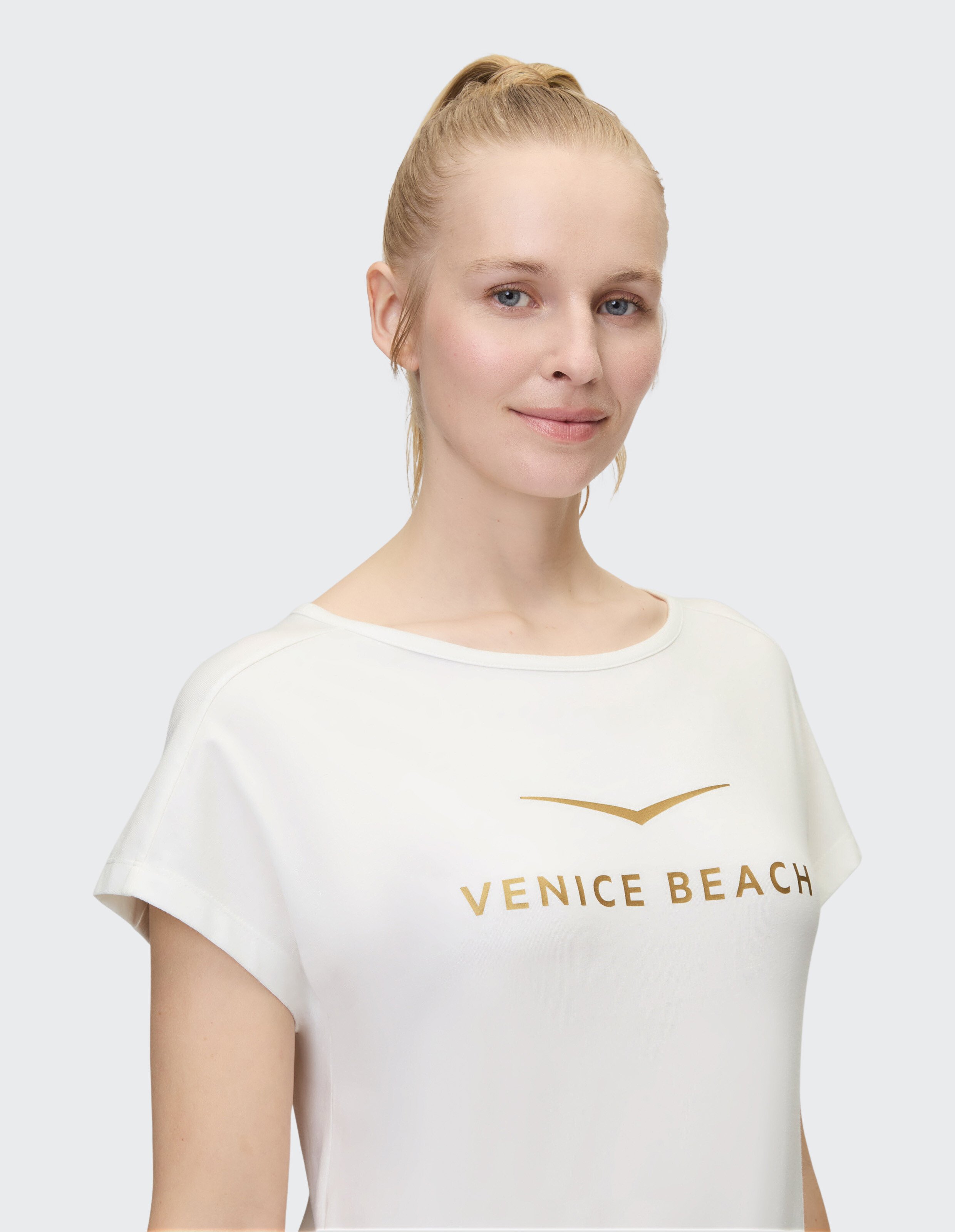 Frau trägt ein weißes Freizeit-T-Shirt mit goldenem „VENICE BEACH“-Aufdruck.
