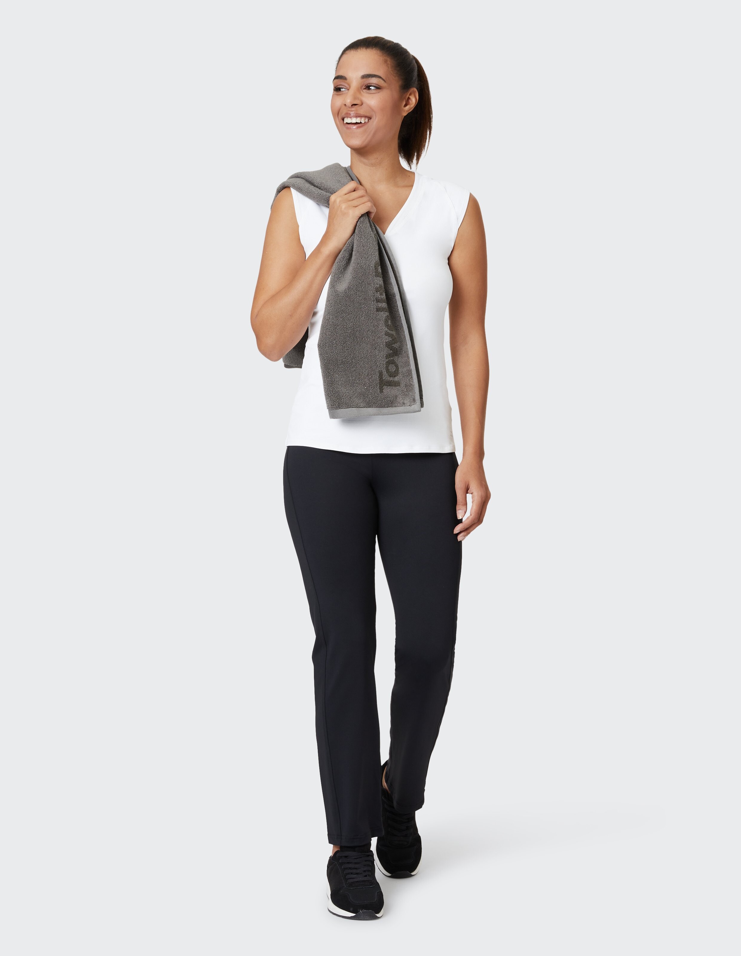 Frau in einem weißen ärmellosen Top und einer schwarzen Hose lächelt mit einem Handtuch.