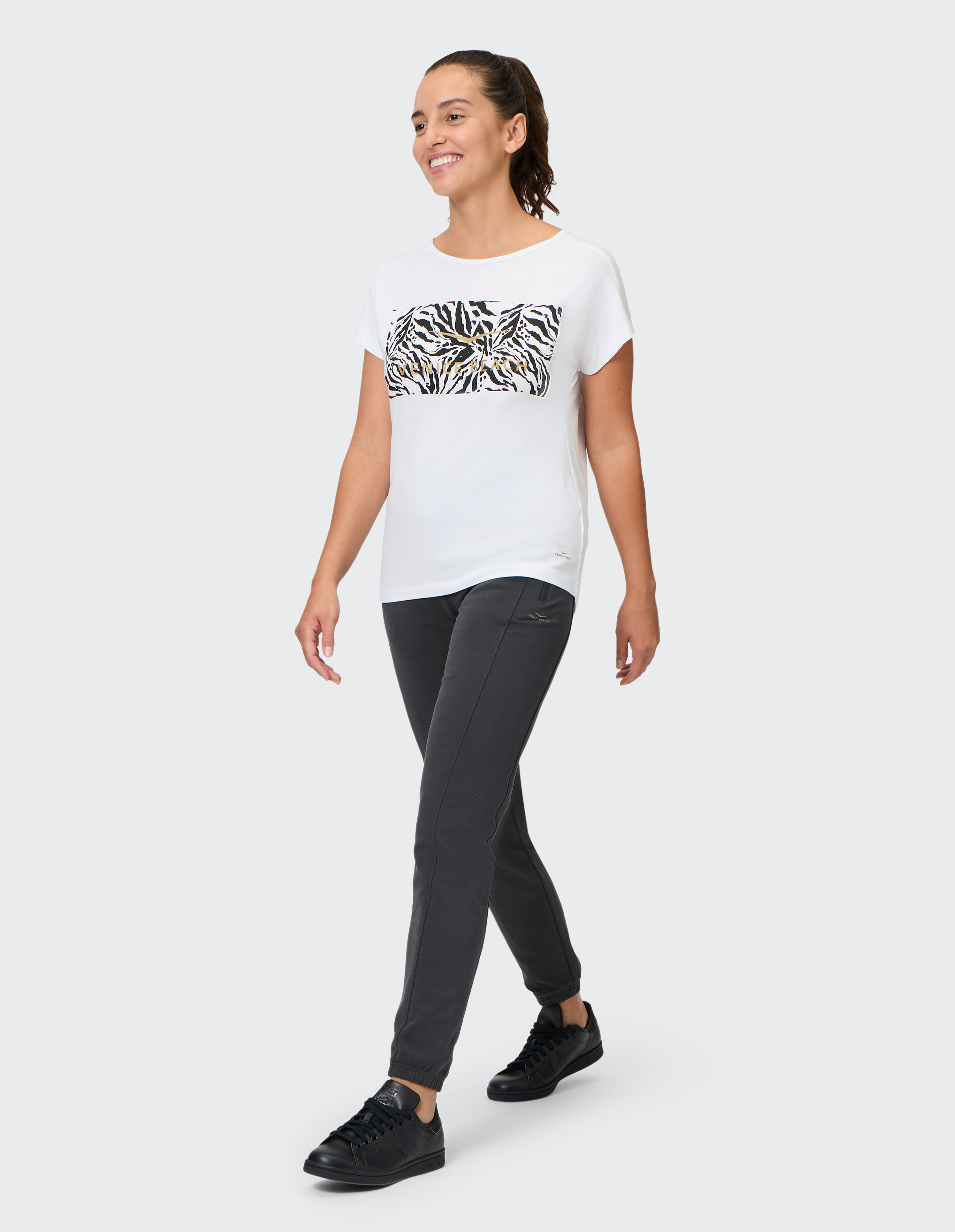 Frau in weißem T-Shirt mit Zebramuster und grauer Hose geht lässig spazieren.