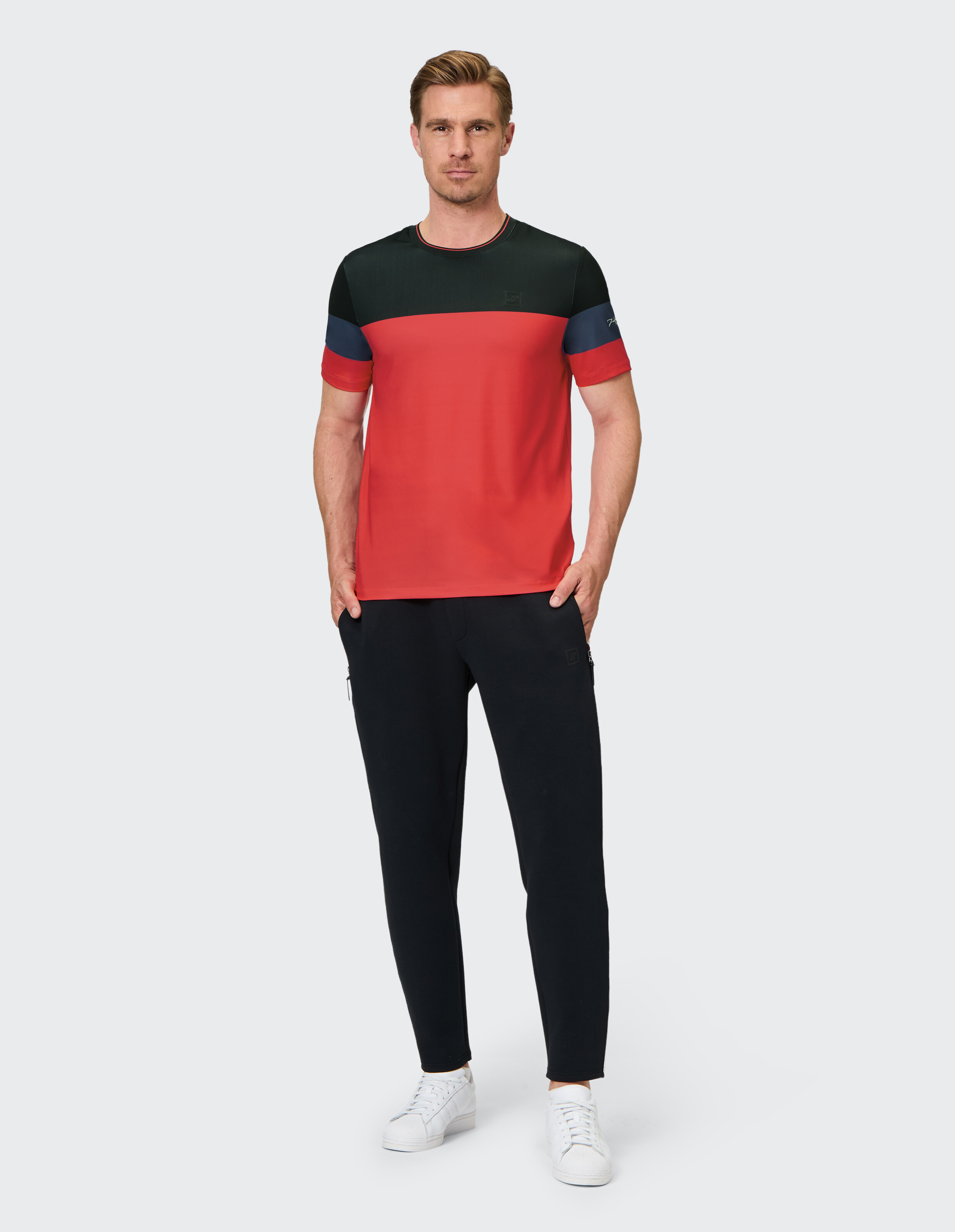Mann trägt ein T-Shirt in den Farben Rot, Schwarz und Blau in Blockfarben und dazu schwarze lässige Jogginghosen.