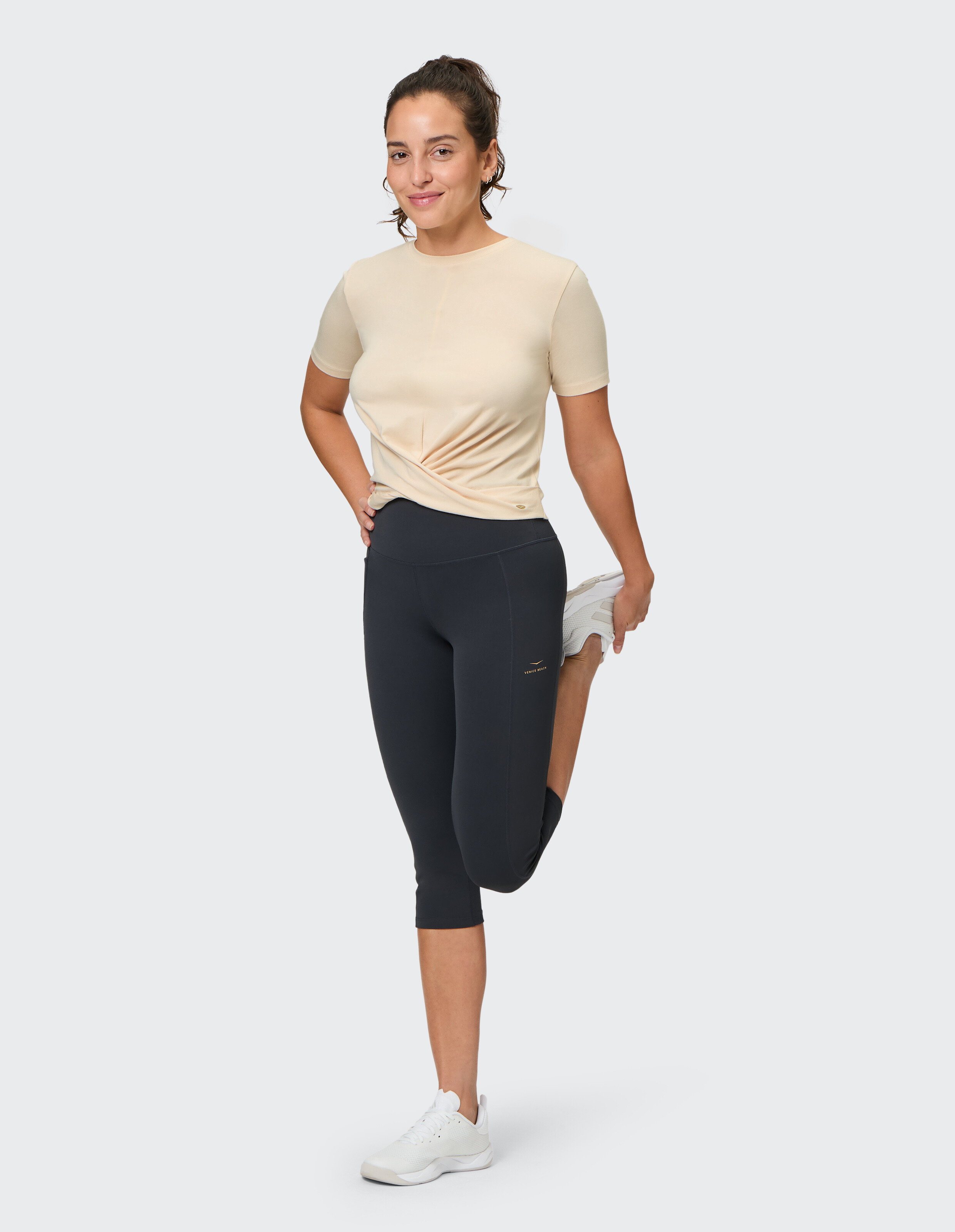 Frau in beigem T-Shirt mit verdrehter Vorderseite und schwarzen Leggings beim Stretching in lässiger Sportkleidung.