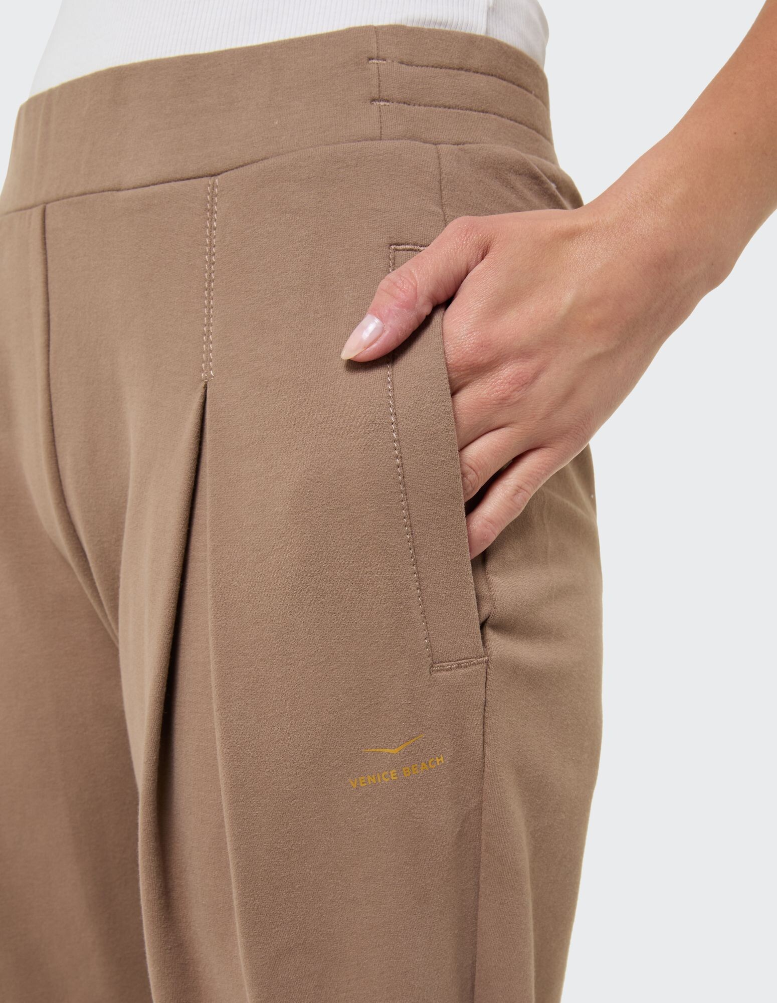 Nahaufnahme einer beigen Freizeithose mit Bundfalten und Seitentaschen und dem Logo von Venice Beach.