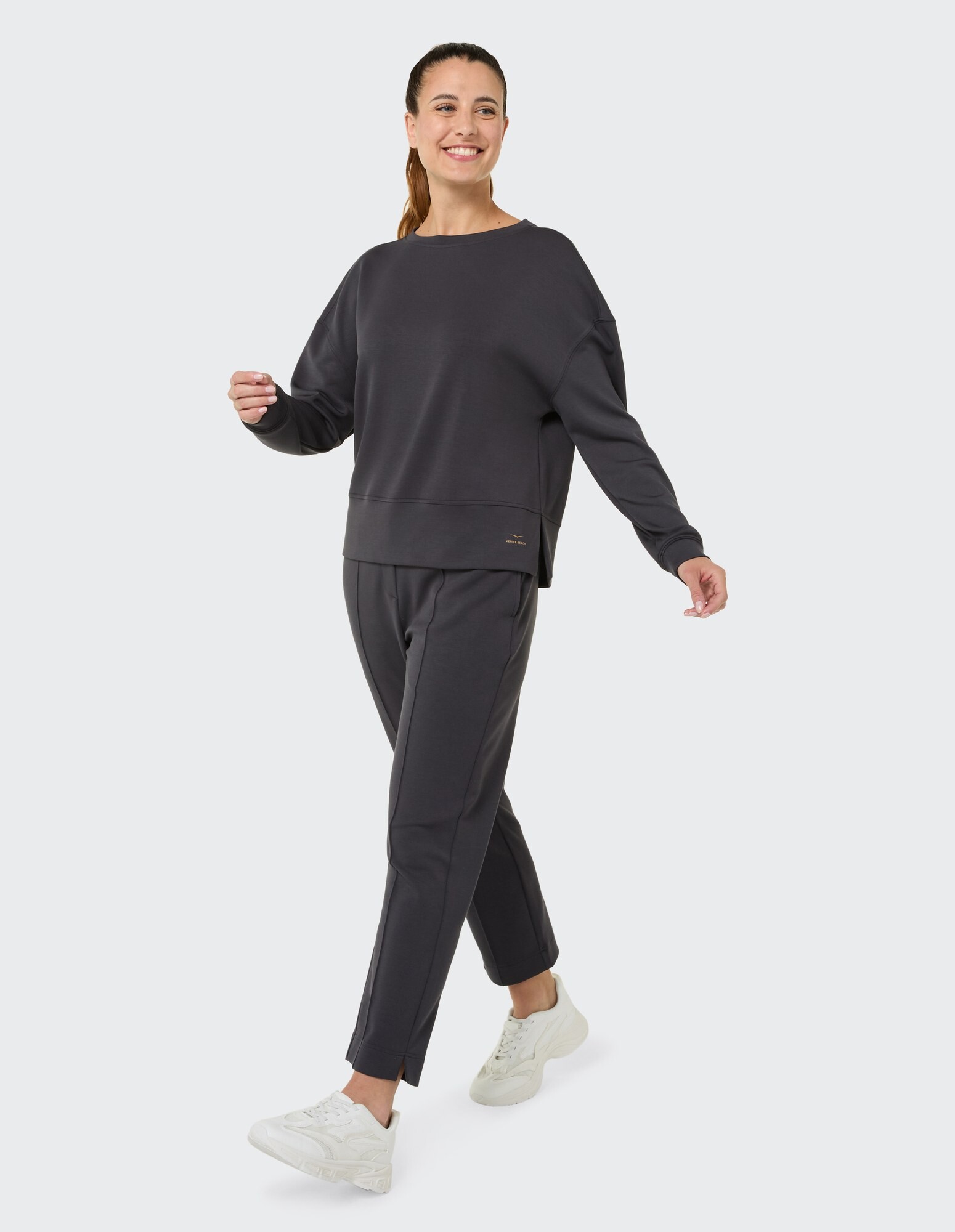 Frau in dunkelgrauem, lässigem Sweatshirt und Hose, lächelnd und gehend.