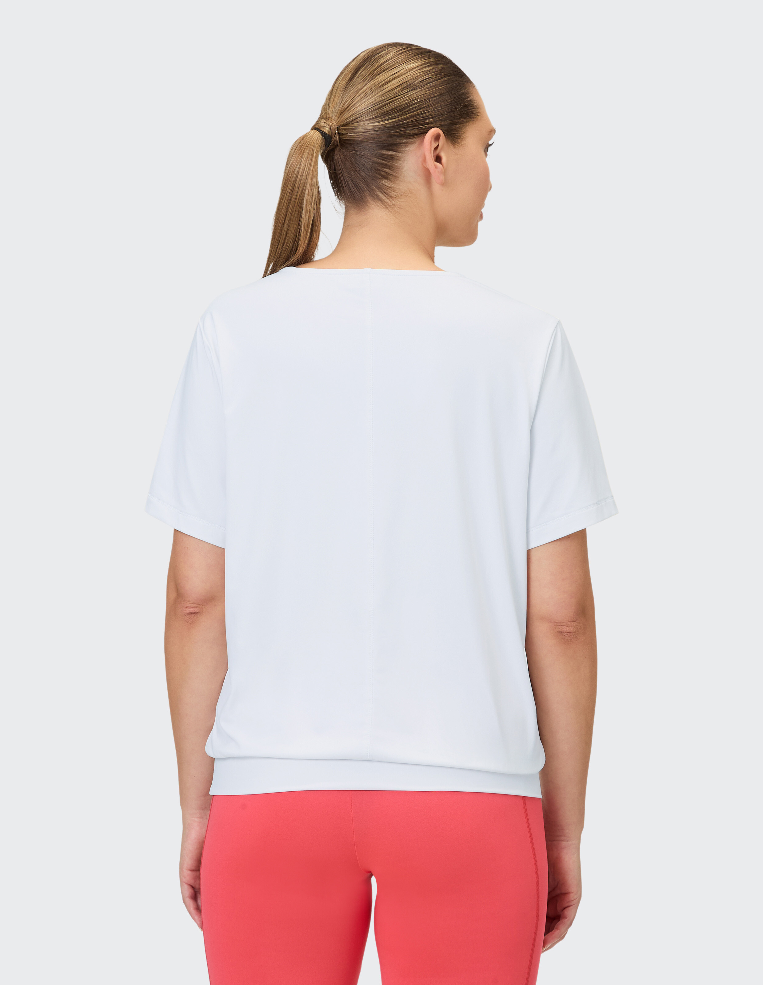 Eine Frau mit Pferdeschwanz trägt ein weißes Kurzarm-Shirt und eine Korallefarbene Leggings.