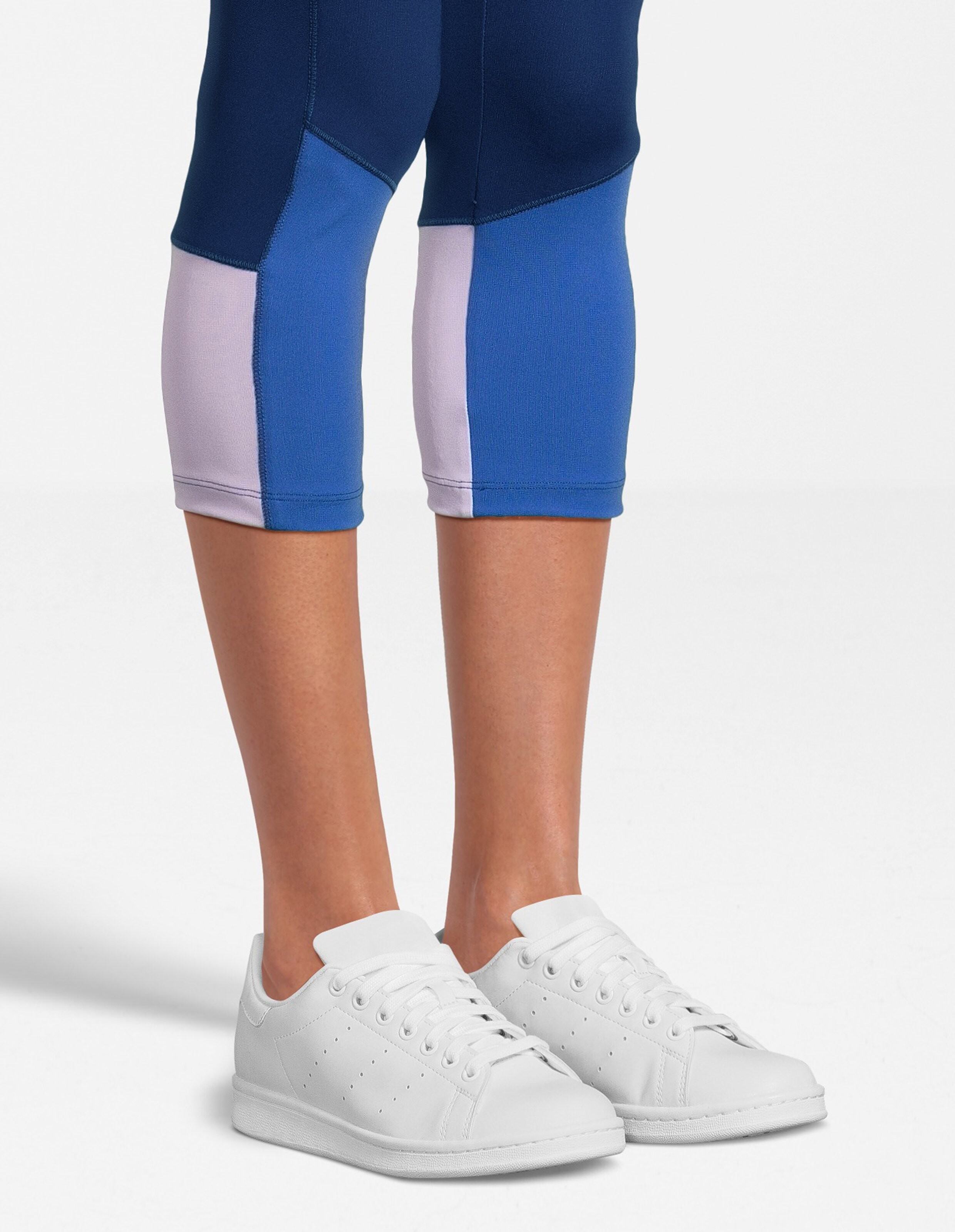 Frau trägt kurze Leggings in Blau und Lila in Colour-Block-Optik und weiße Turnschuhe.