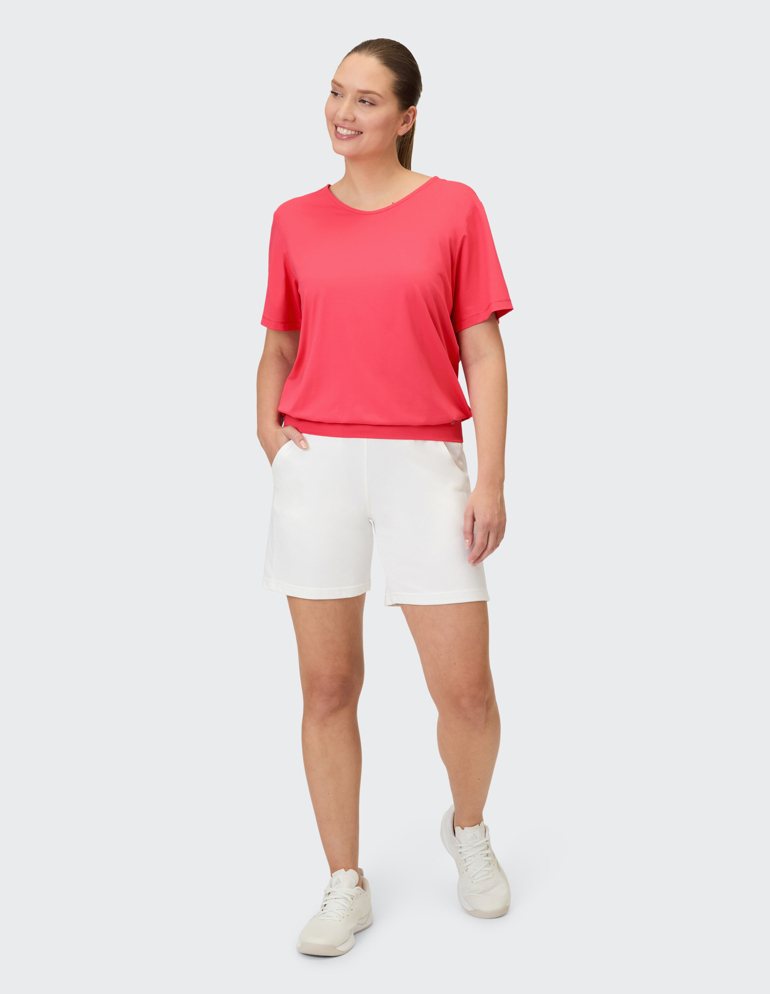 Frau im leuchtend korallenroten T-Shirt und weißen Shorts mit Sneakern.
