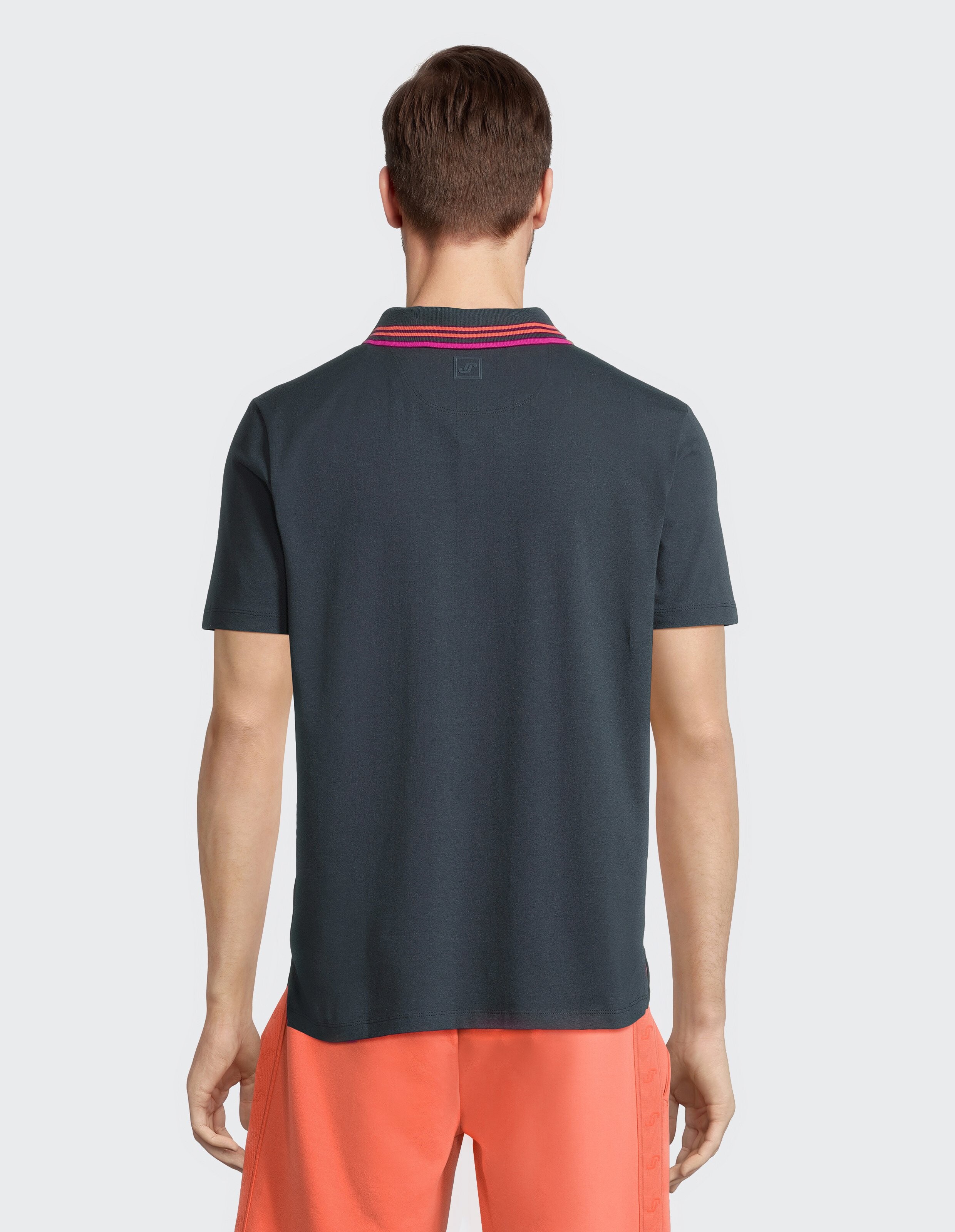 Mann in dunkelblauem Poloshirt mit rosa Kragenstreifen und roten Shorts, von hinten gesehen vor einem hellgrauen Hintergrund.