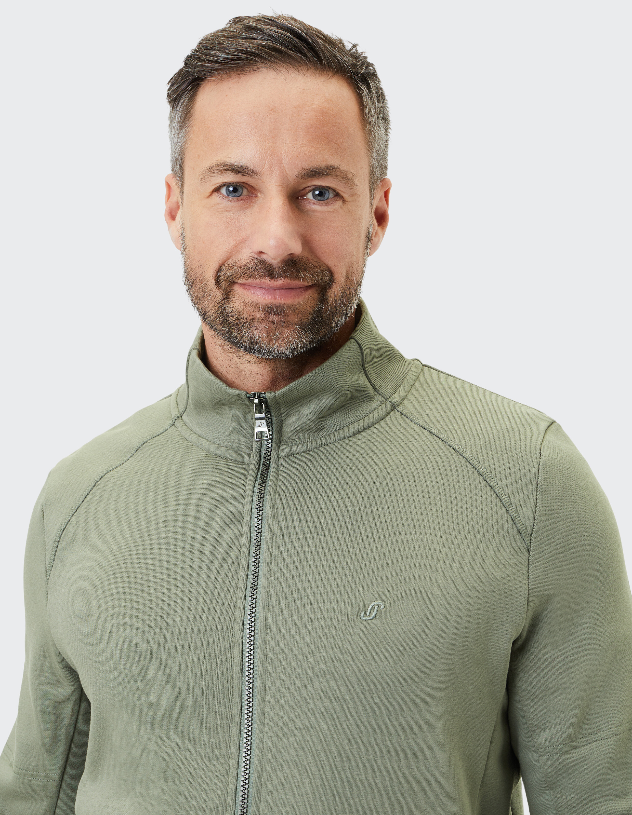 Mann mit grauem Haar und Bart lächelt in die Kamera und trägt eine hellgrüne Jacke mit Reißverschluss auf grauem Hintergrund.