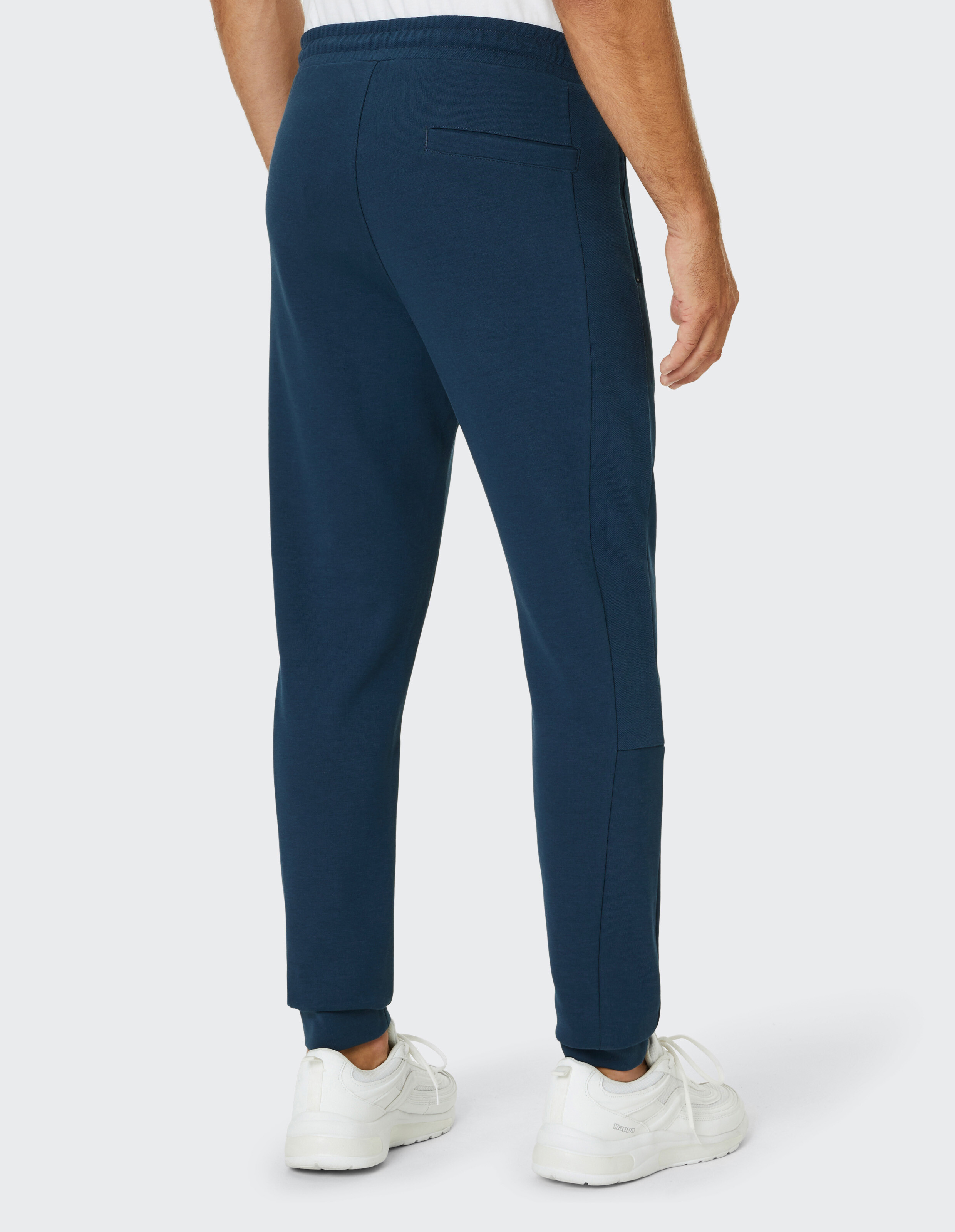 Mann in blauer Jogginghose und weißen Turnschuhen, von hinten zu sehen, trägt ein weißes Shirt, Hände locker an den Seiten.