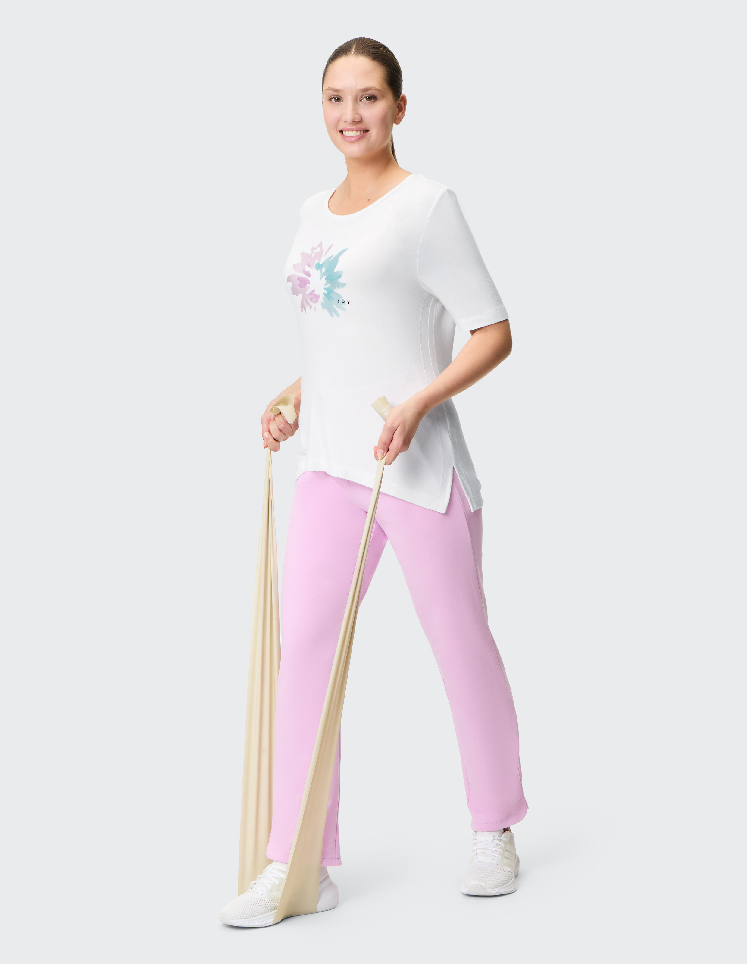 Frau in einem weißen Grafik-T-Shirt und einer rosafarbenen Lounge-Hose trainiert mit einem Widerstandsband.