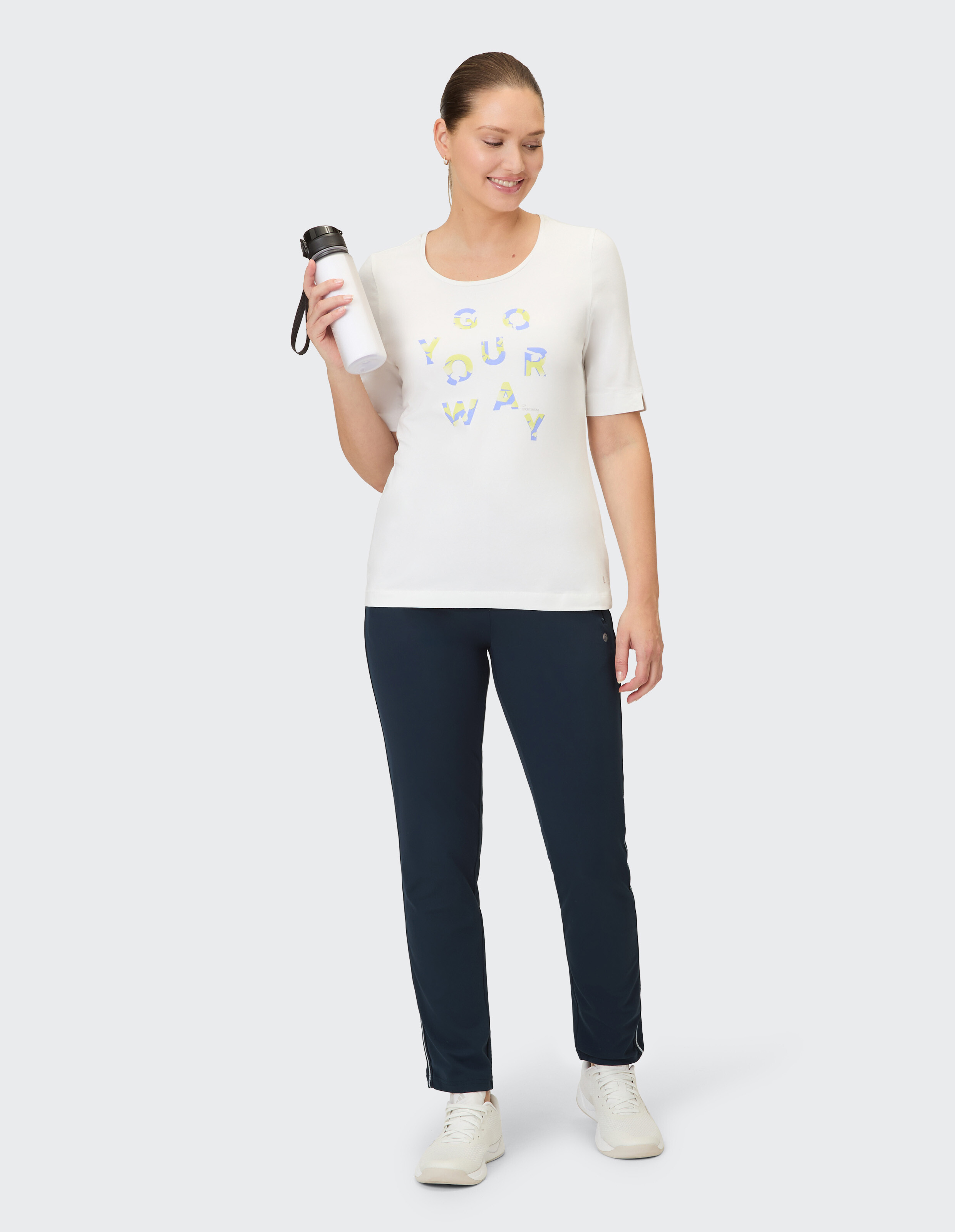 Frau in weißem Grafik-T-Shirt und dunkelblauer Hose, hält eine Wasserflasche in der Hand und lächelt.
