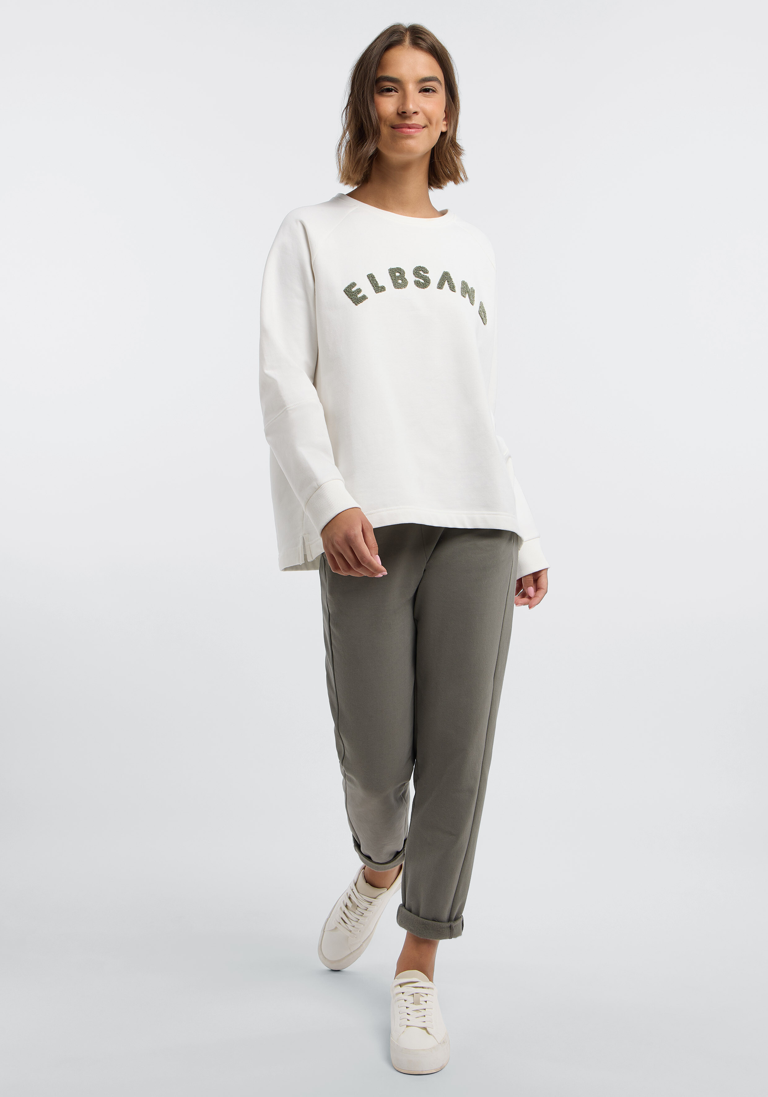 Frau trägt ein weißes ELBSAND-Sweatshirt und eine olivgrüne Freizeithose.