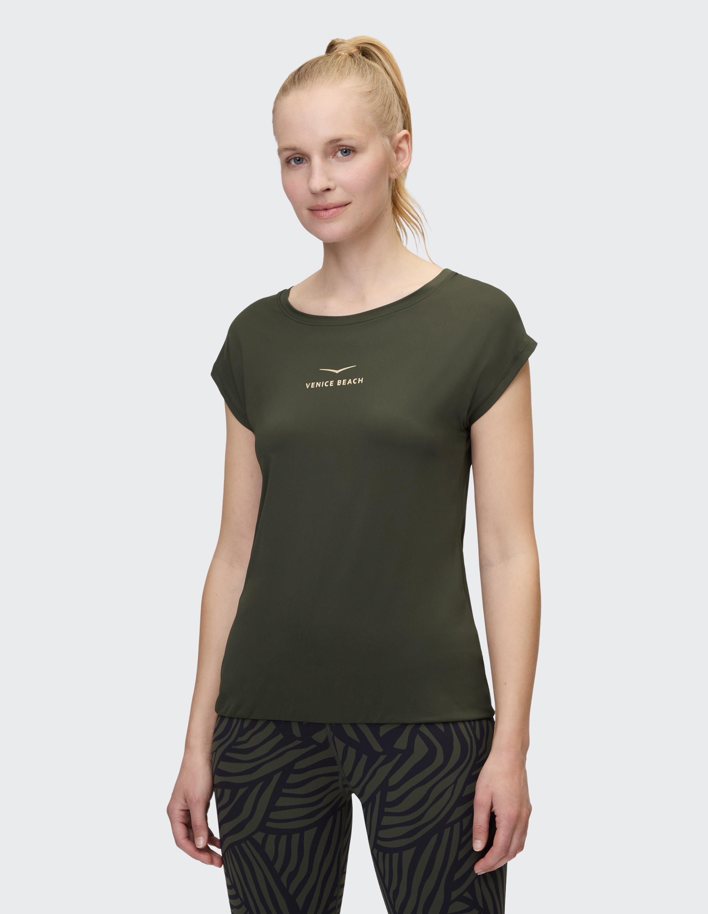 Frau in olivgrünem T-Shirt mit Venice Beach-Logo, gepaart mit gemusterten Leggings.