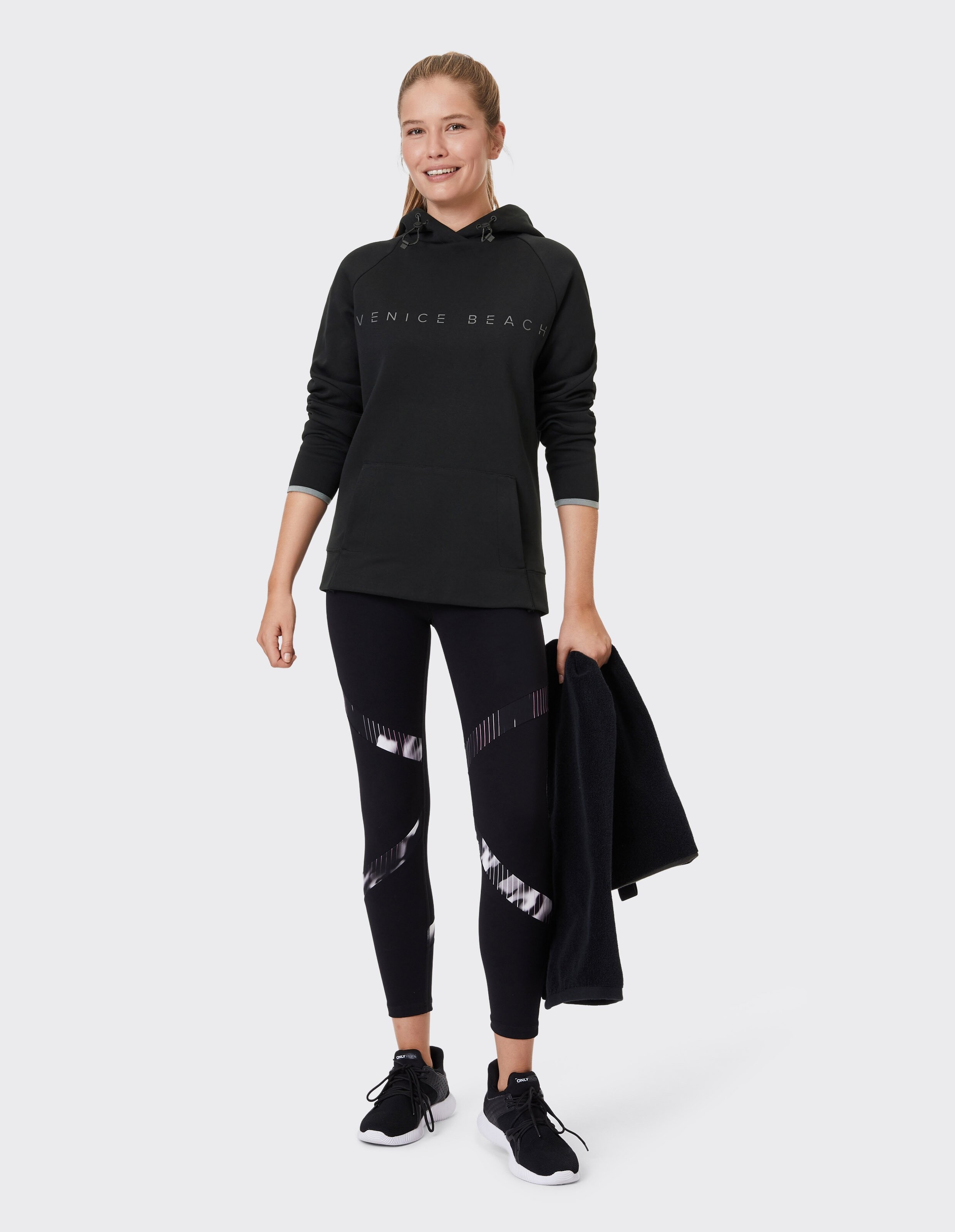 Frau trägt einen schwarzen Kapuzenpullover von Venice Beach und gemusterte Leggings für gemütliche Freizeitkleidung.