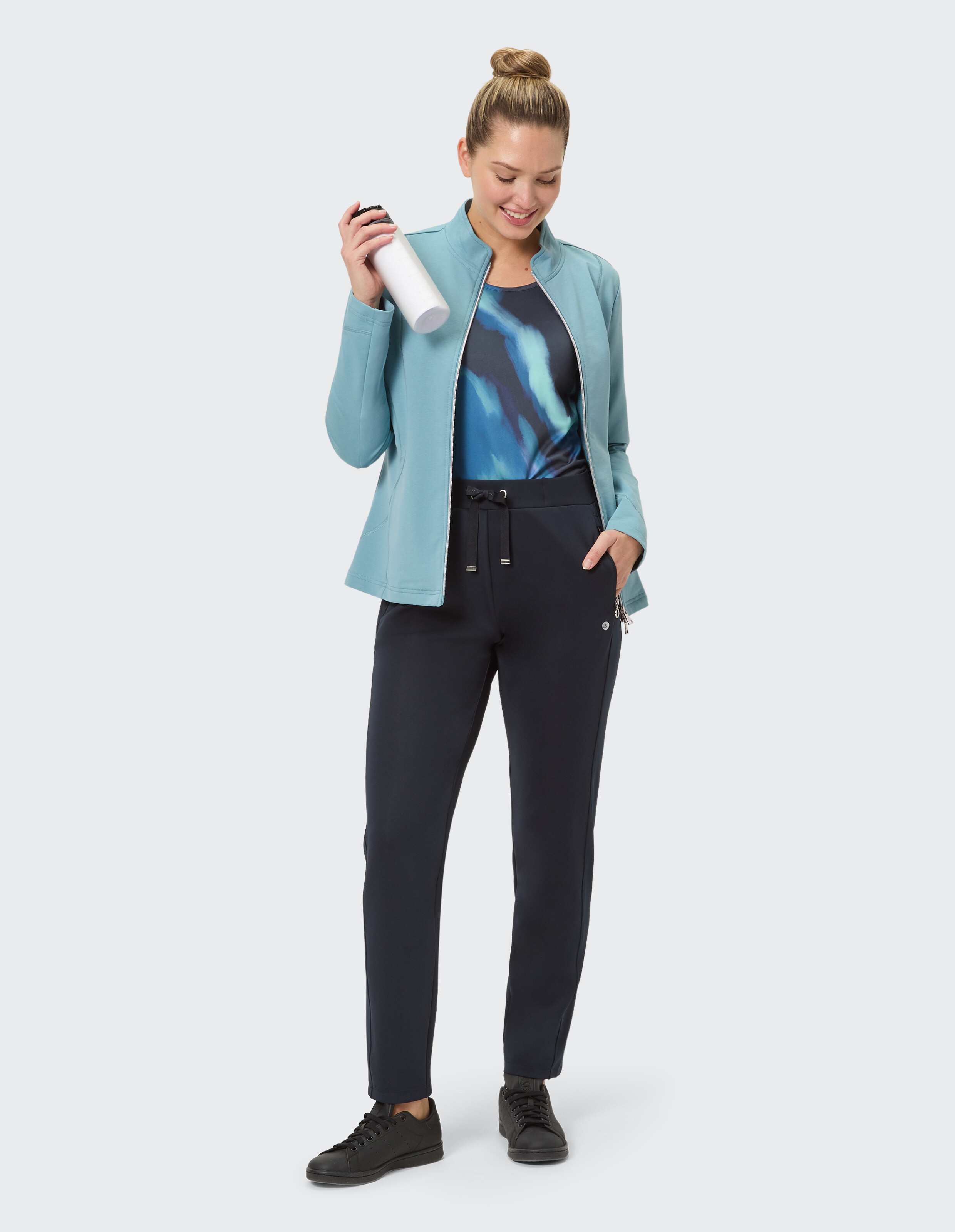 Frau in hellblauer Jacke und dunkelblauer Hose, eine Wasserflasche in der Hand, lässiger Stil.