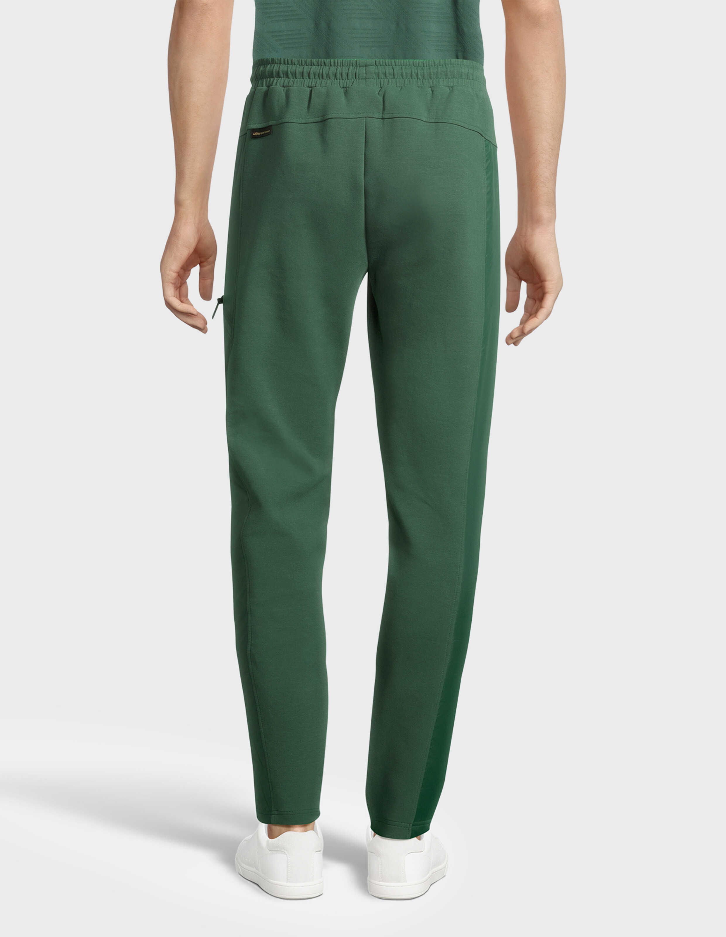 Eine Person trägt eine grüne Jogginghose und weiße Sneaker, von hinten zu sehen, vor einem neutralen, grauen Hintergrund.