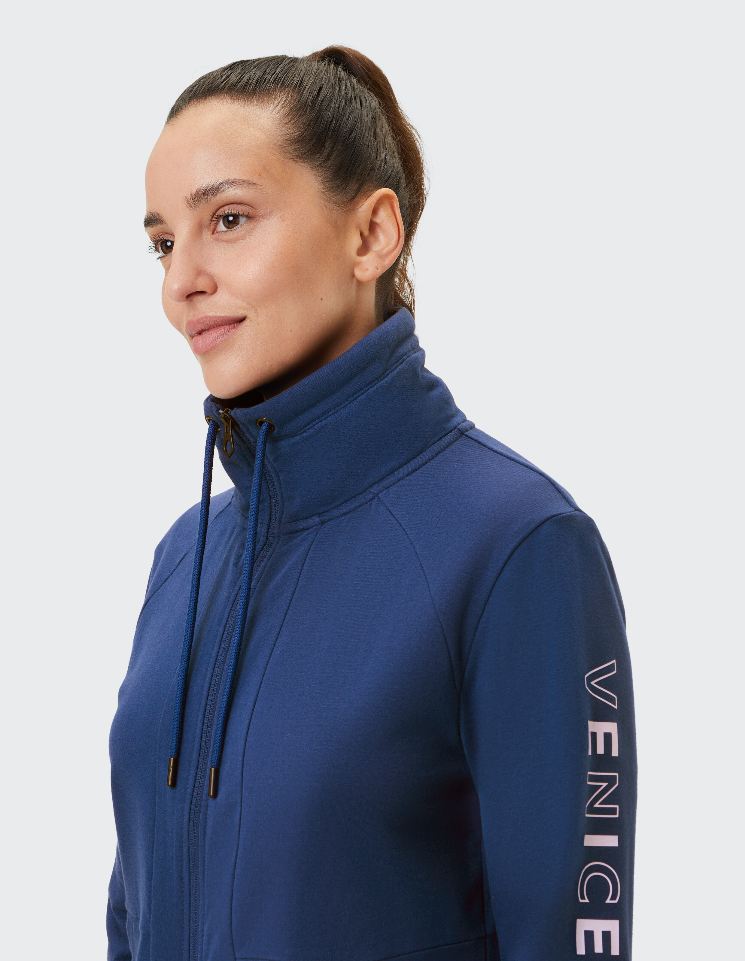 Nahaufnahme einer Frau in einer dunkelblauen Jacke mit Reißverschluss, hohem Kragen und „VENICE“-Aufdruck auf dem Ärmel, ideal für Freizeitkleidung.