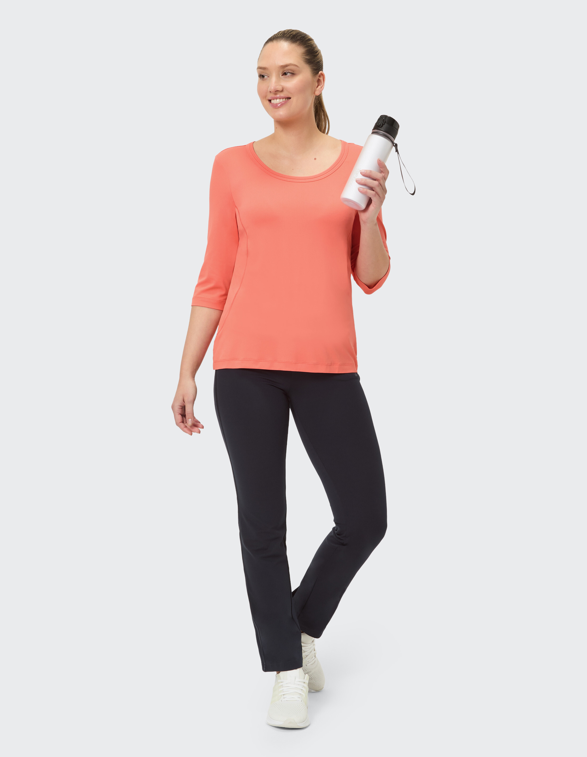 Frau in einem korallenroten Top und einer schwarzen Hose, eine Wasserflasche in der Hand, lässiger Look.