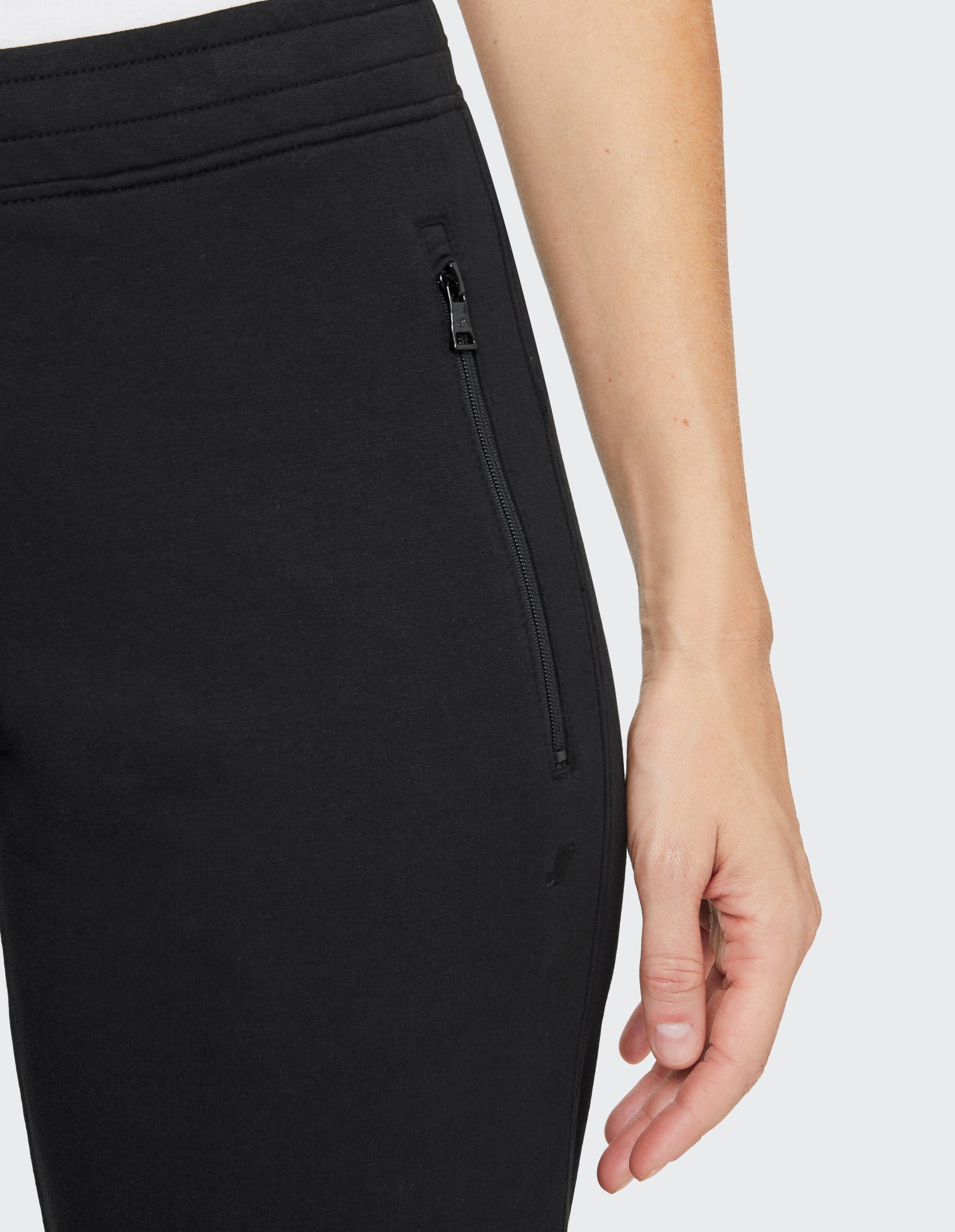 Ein Arm ruht locker neben einer schwarzen Jogginghose mit Reißverschlusstasche, vor hellem Hintergrund.