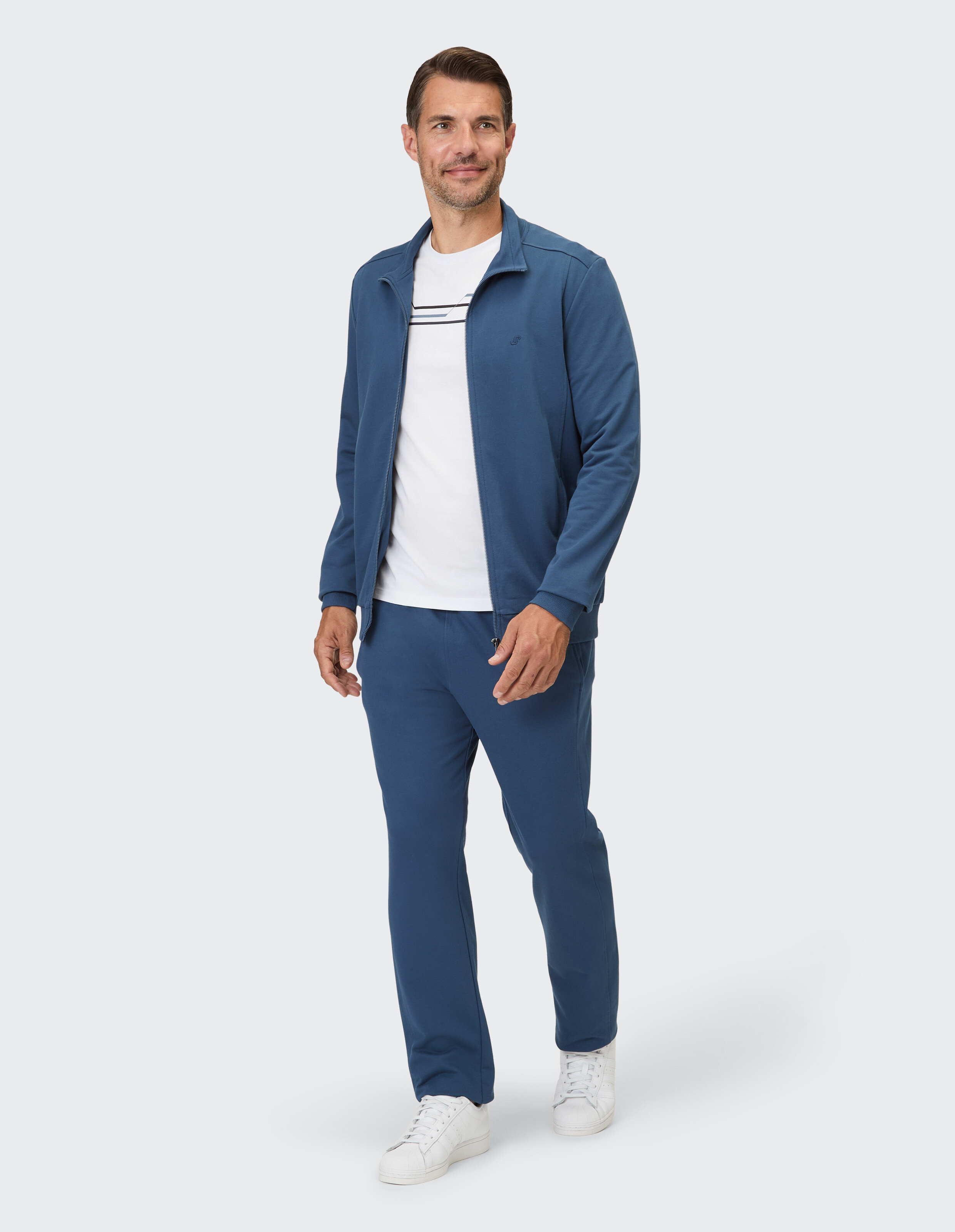 Ein Mann in einem blauen Trainingsanzug mit Reißverschluss, passender Hose und weißem T-Shirt mit Streifen.